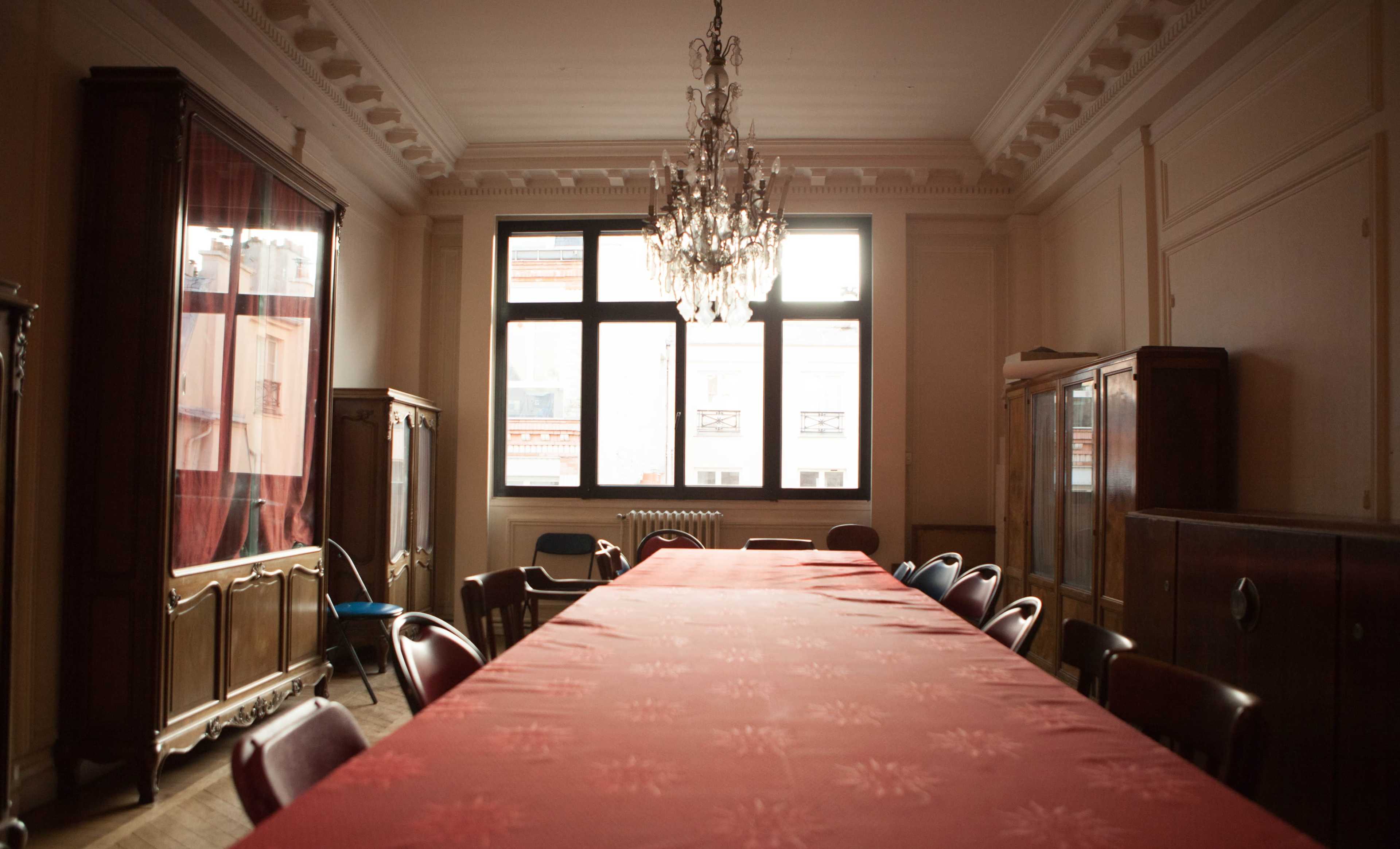 A long table covered with a red tablecloth is set in a well-lit room featuring a chandelier and wooden furniture.