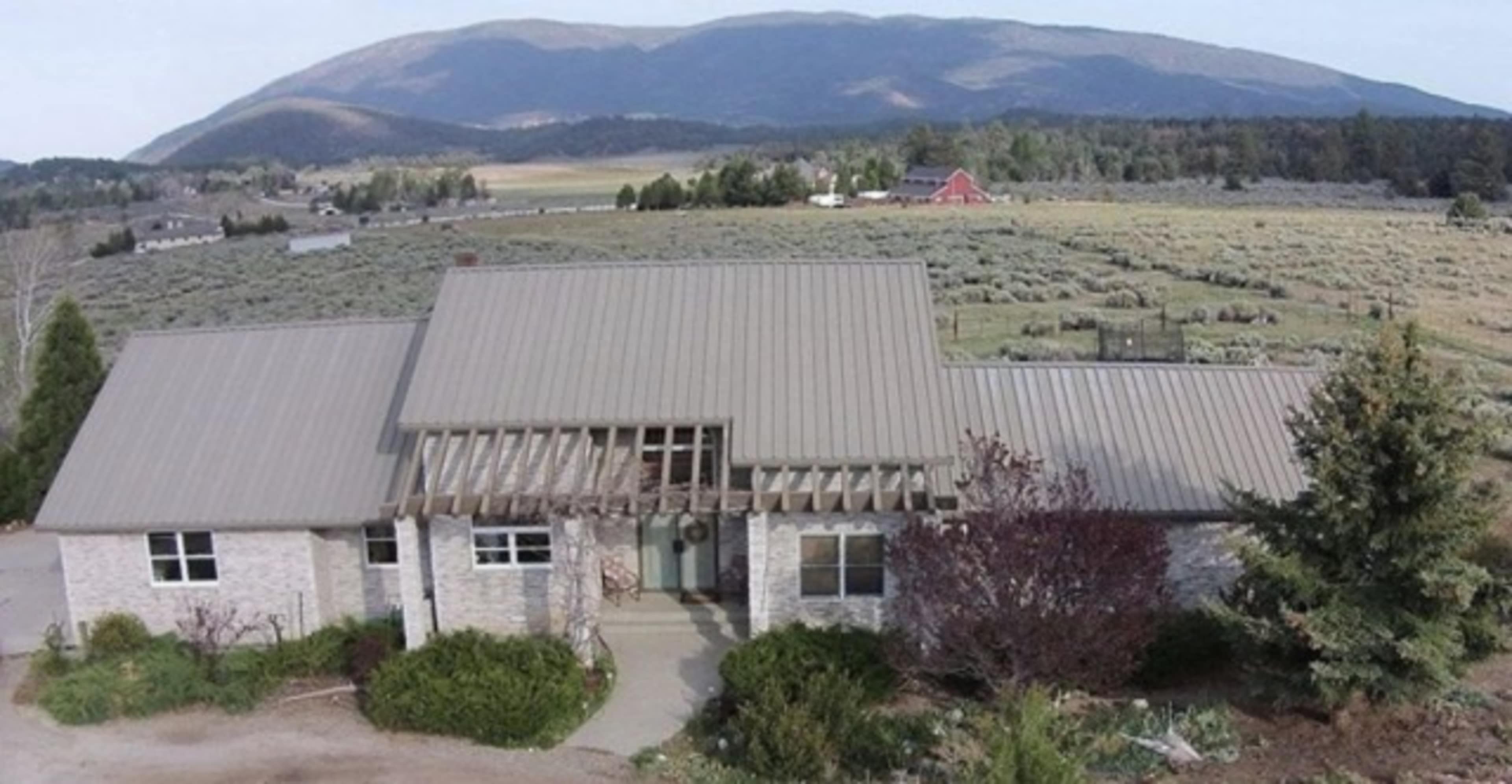 A single-story house with a metal roof, set against a backdrop of mountains and fields with a few trees and a red barn in the distance.