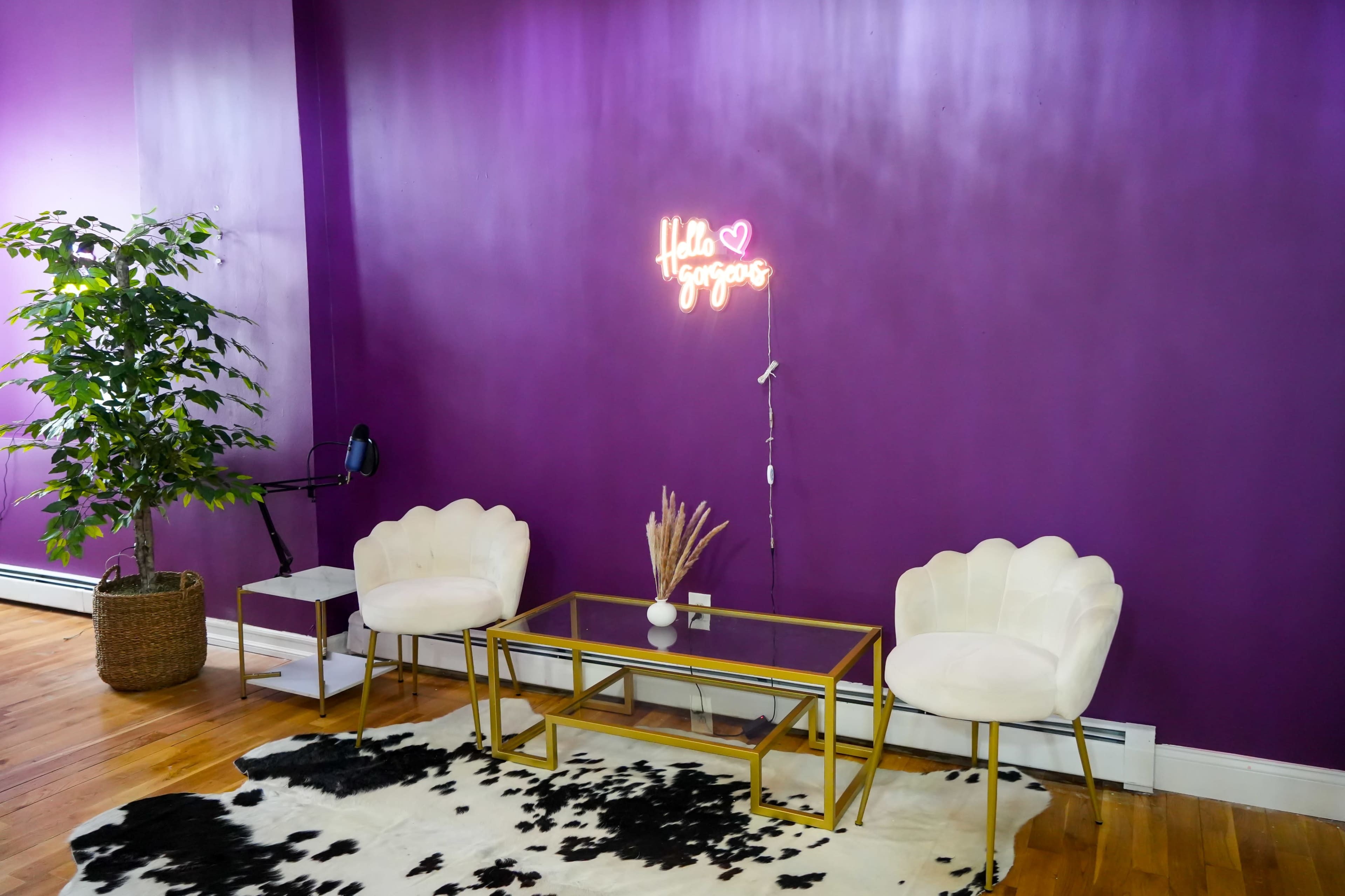 A seating area with two white chairs and a glass table on a black and white cowhide rug, beneath a purple wall featuring a neon sign that says "Hello Gorgeous."