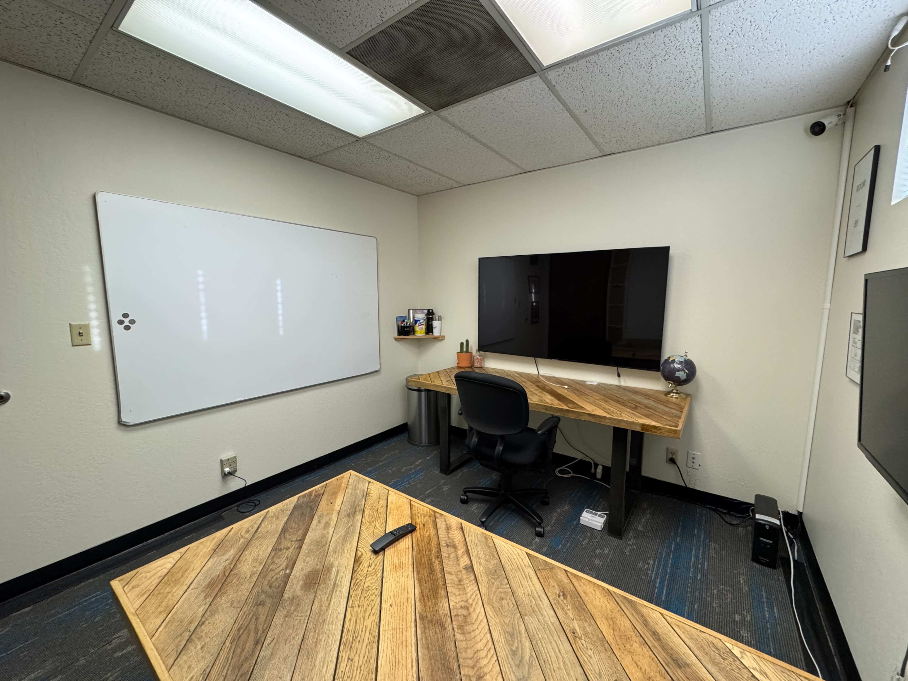 The image shows a simple office space featuring a wooden desk with a computer monitor, a whiteboard on one wall, and a chair on carpeted flooring.