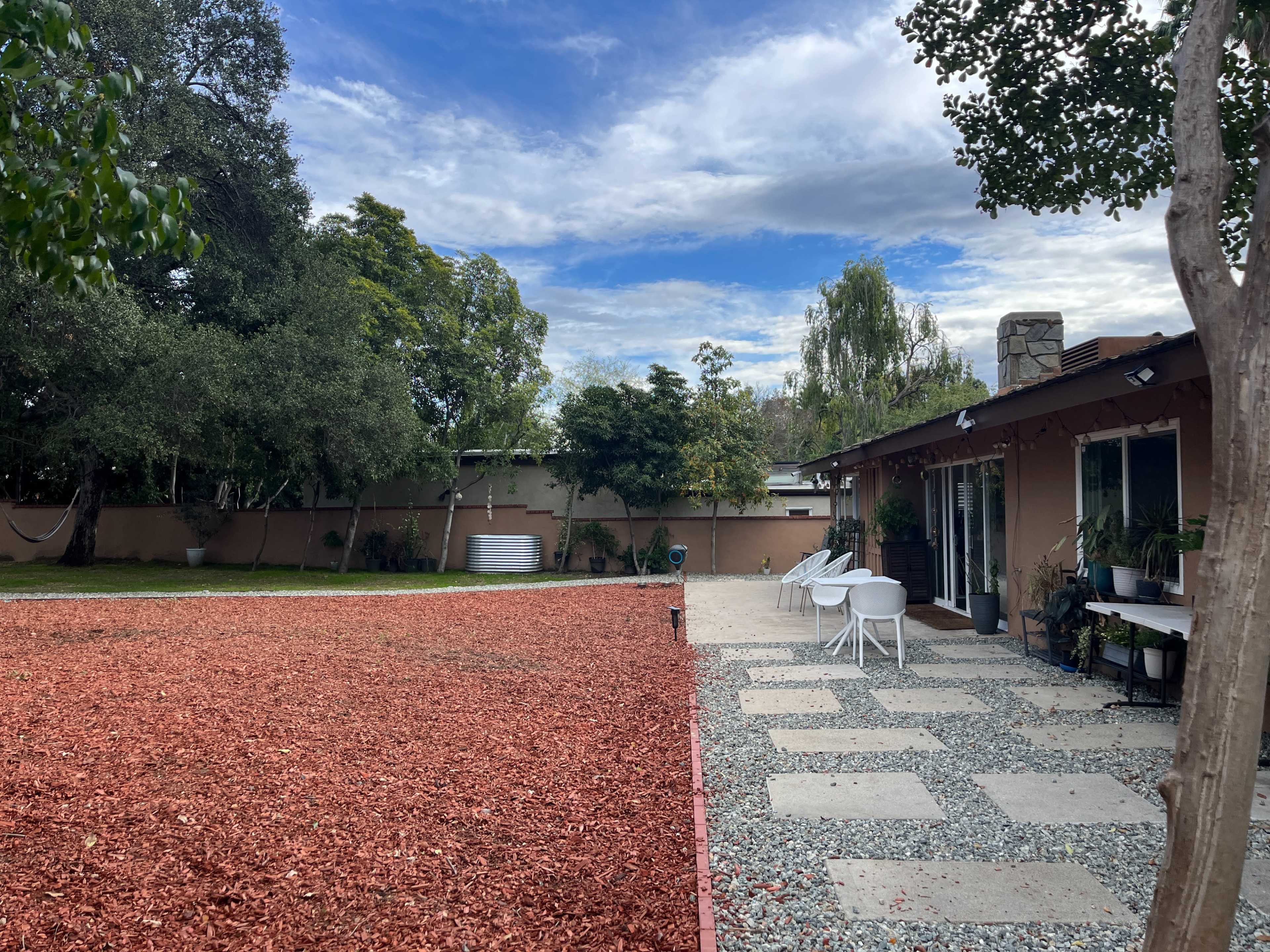A patio area with stone pathways and white chairs is surrounded by a red mulch-covered yard and trees under a cloudy sky.