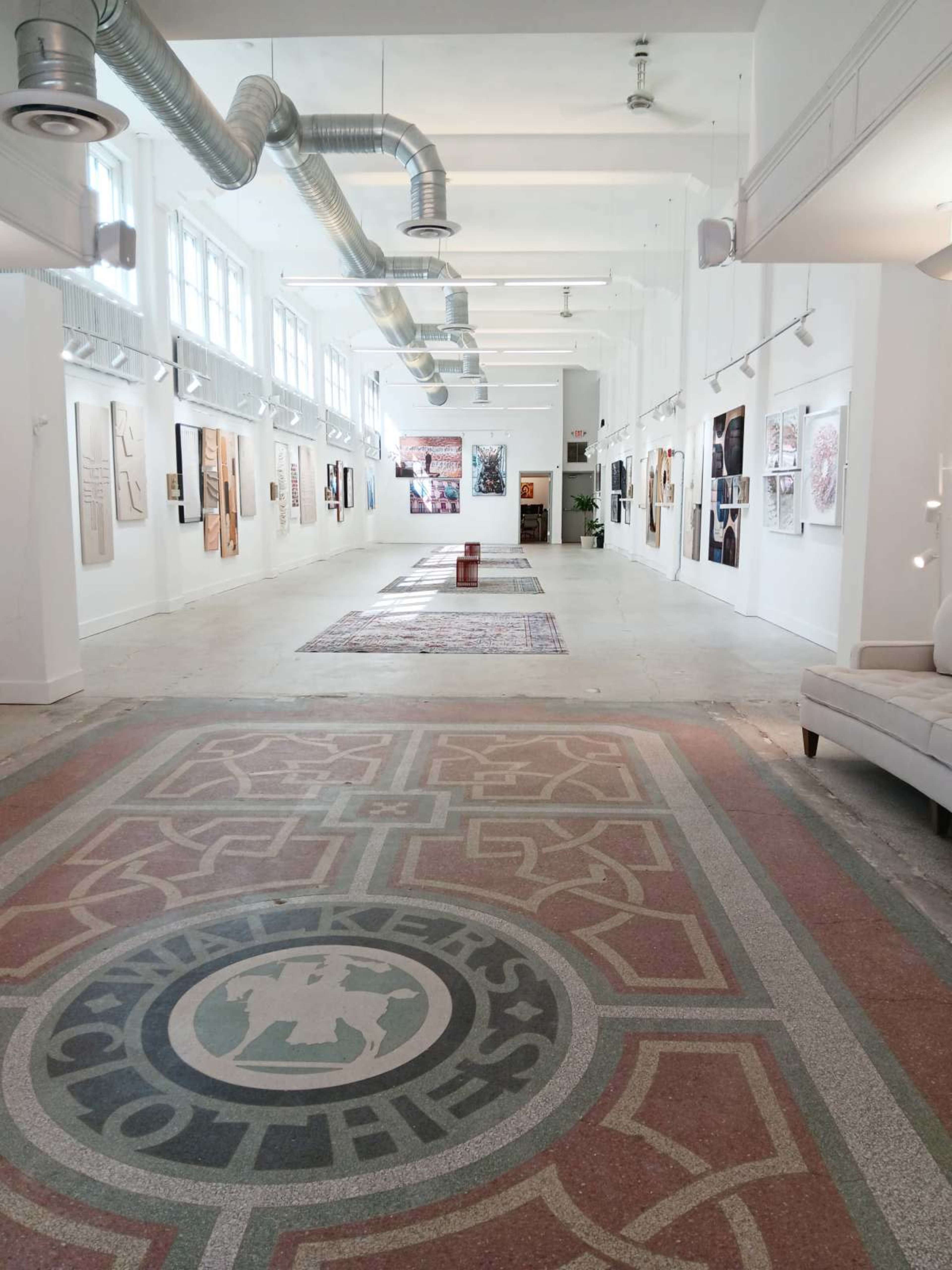 The image shows a spacious art gallery with white walls displaying various artworks and a patterned floor.