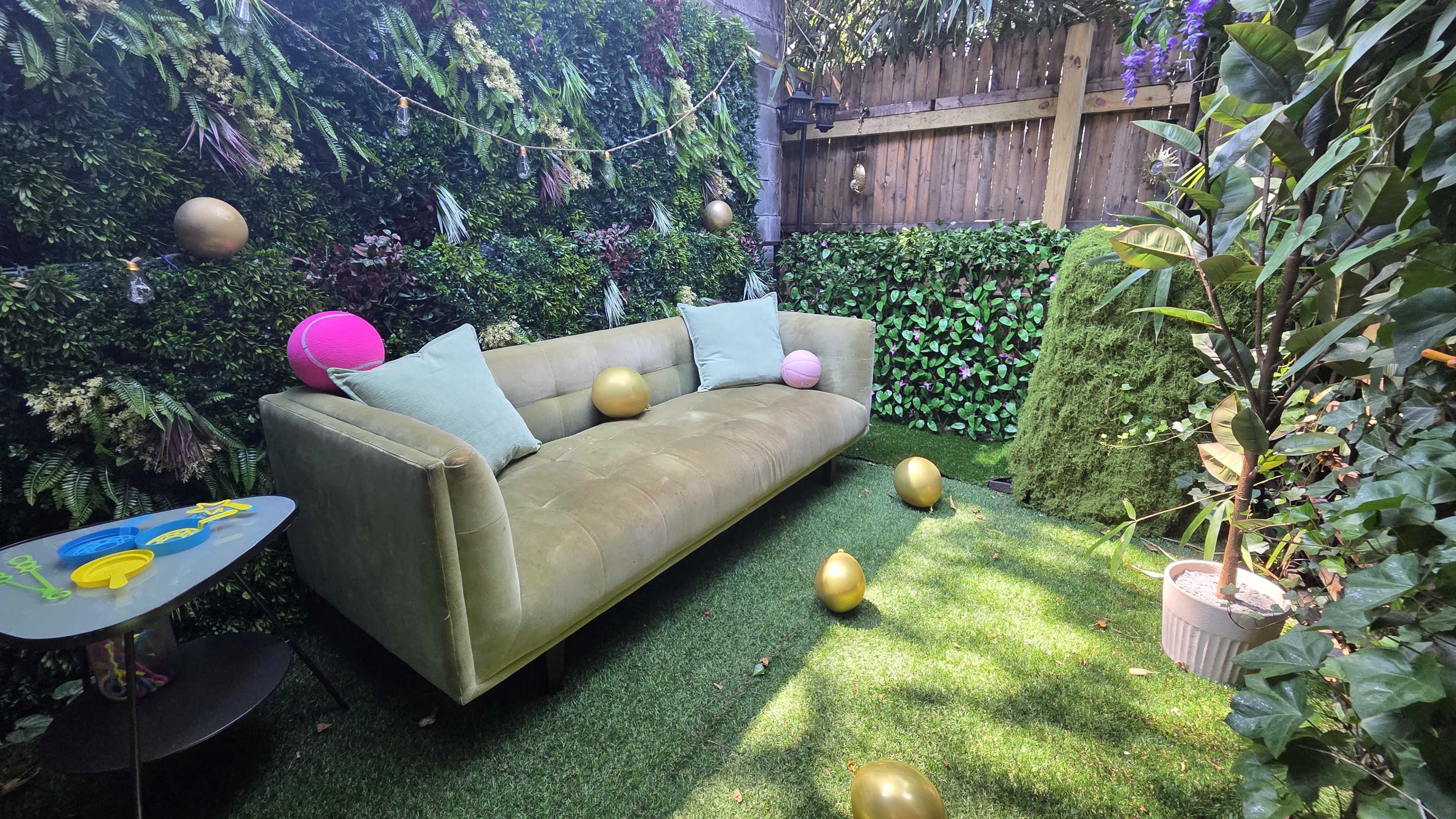 The image shows a green, lush garden area featuring a pale green couch, decorative pillows, and several round golden balls scattered on the grass.