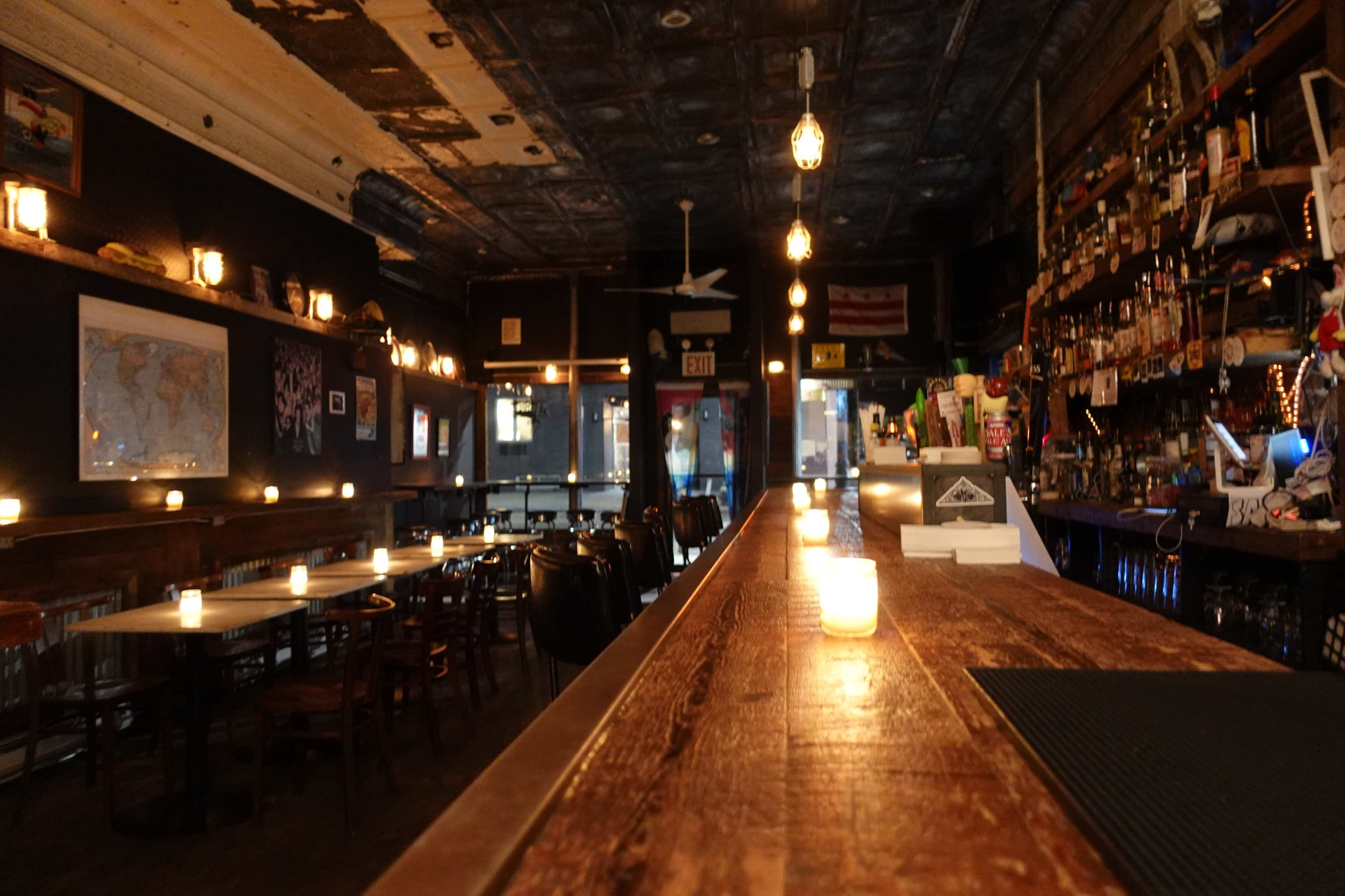 A dimly lit bar with wooden tables and a long counter, adorned with hanging lights and a wall lined with shelves of bottles.