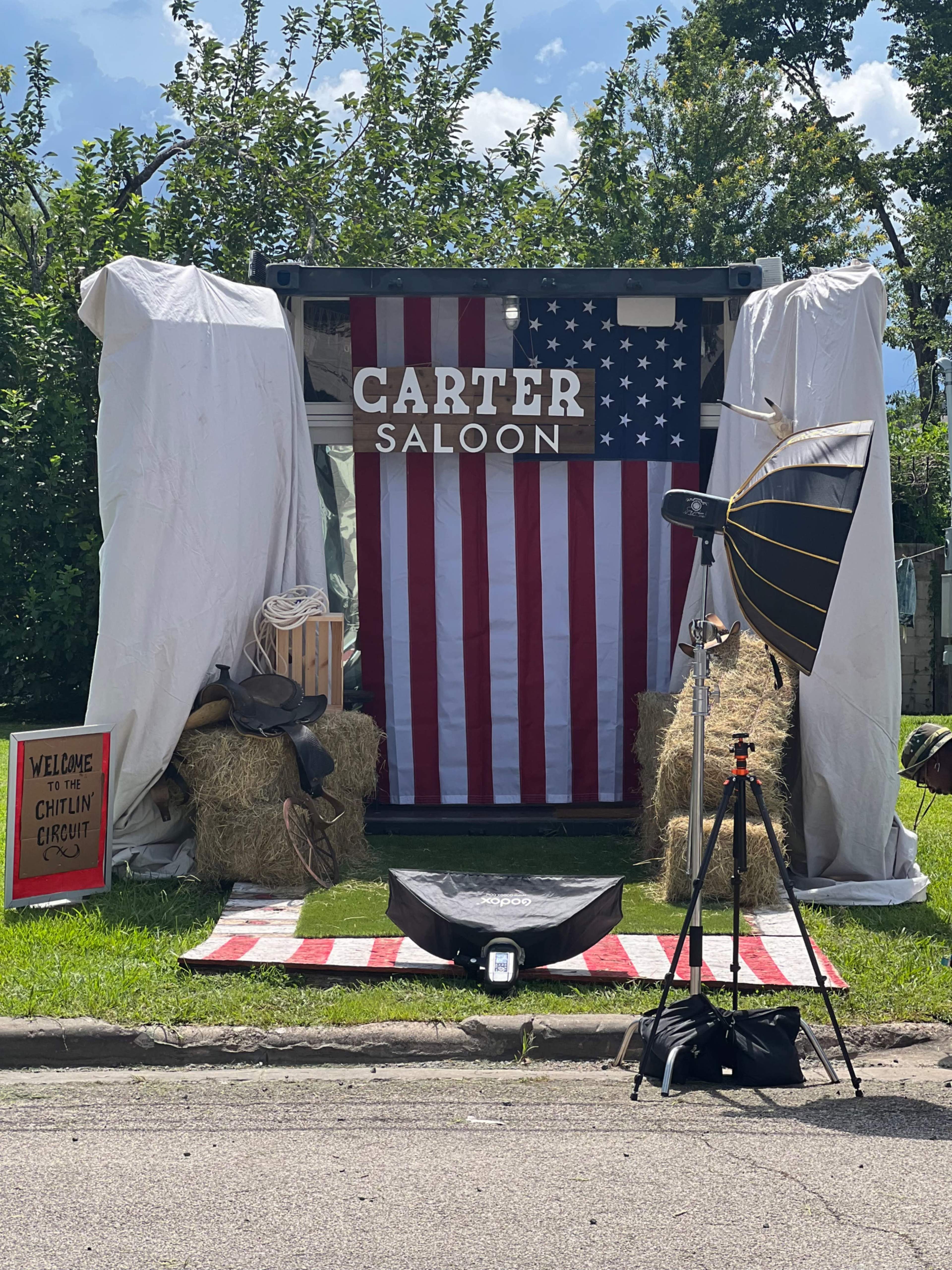 The scene features a saloon-themed setup with a large American flag backdrop, hay bales, and a photography lighting setup on a grassy area.