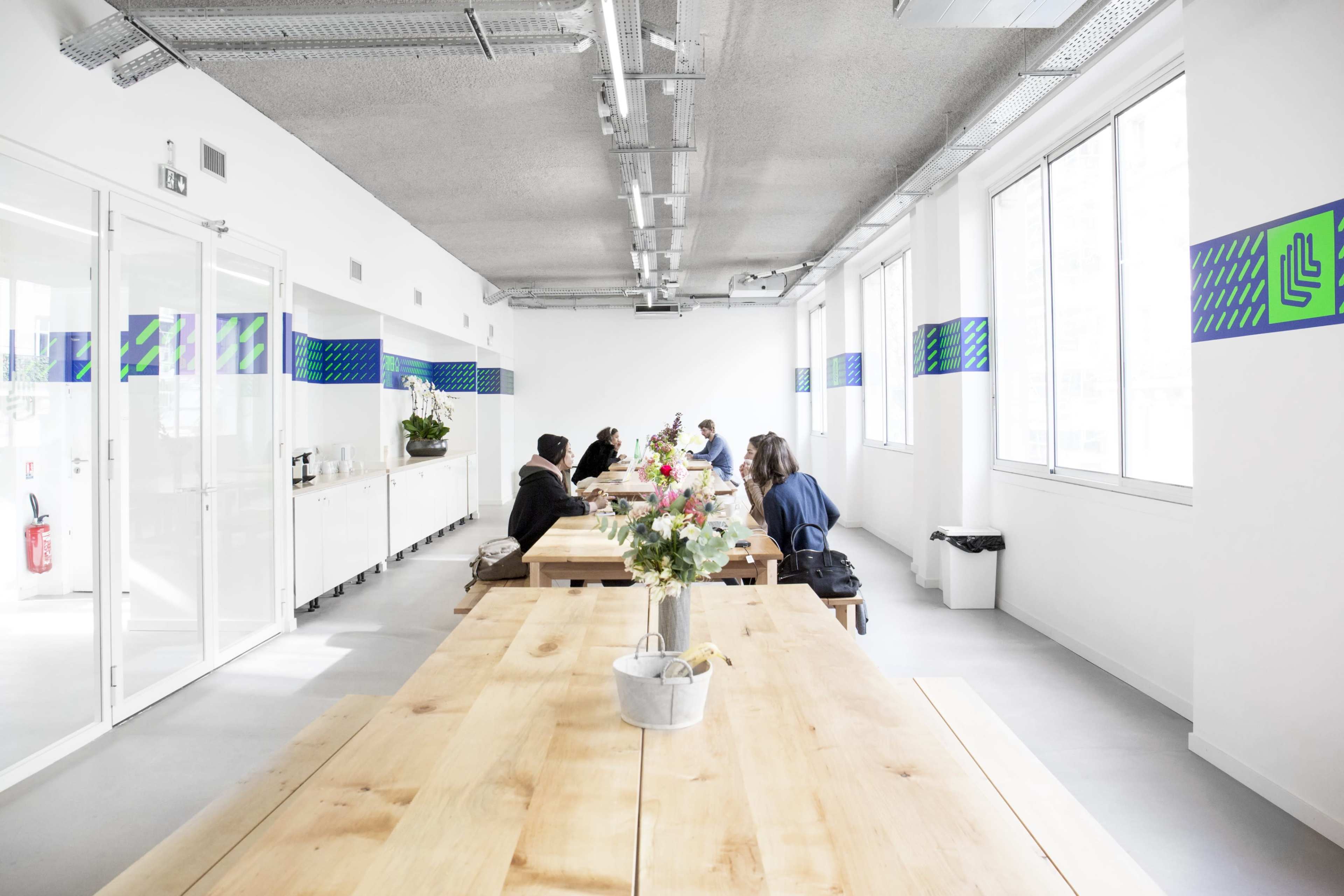 A long wooden table is surrounded by several people in a bright, modern office space with large windows and decorative wall designs.