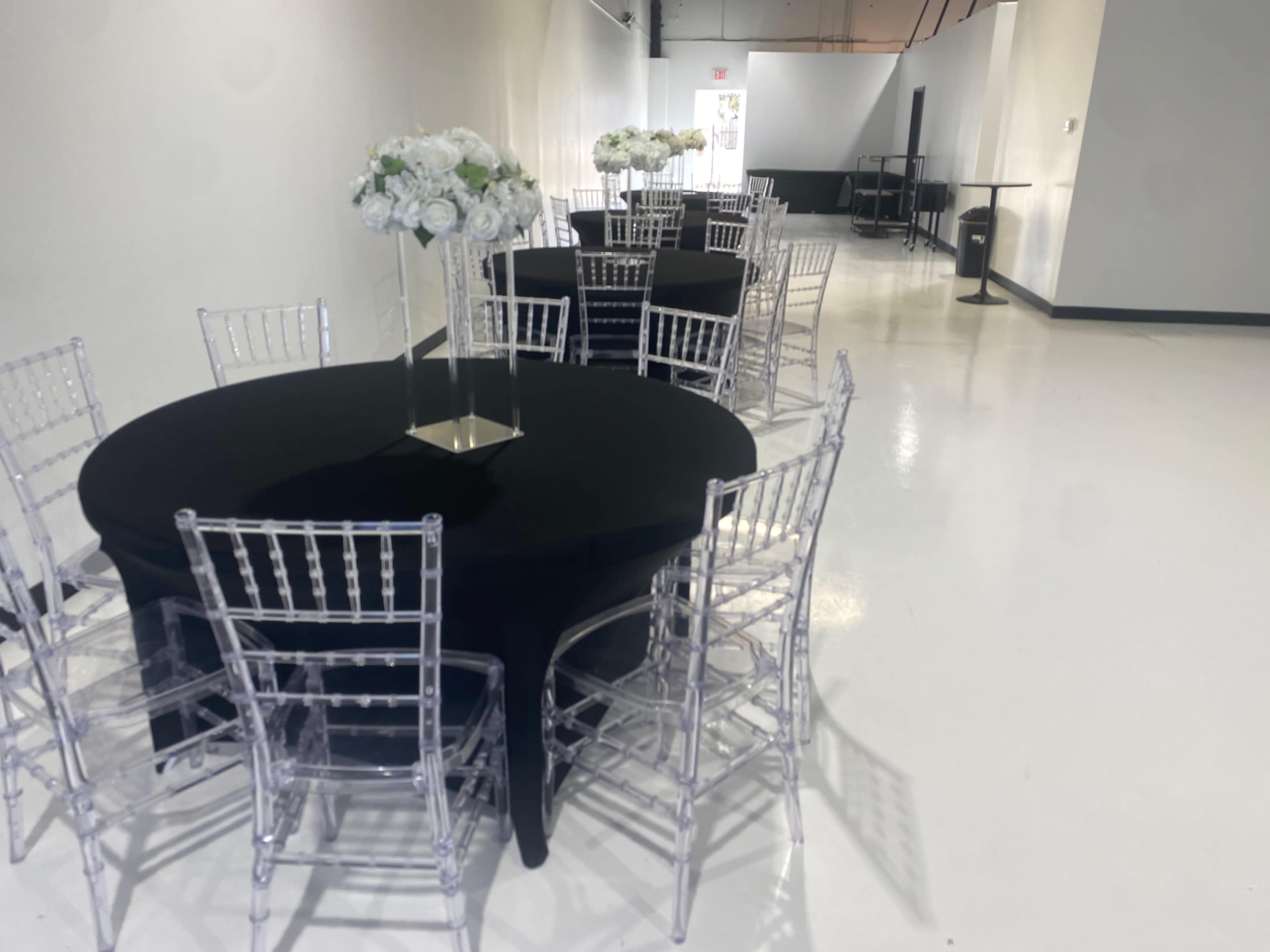 The image shows a clean, modern event space with black tablecloths on round tables and transparent chiavari chairs, adorned with centerpieces of white flowers.