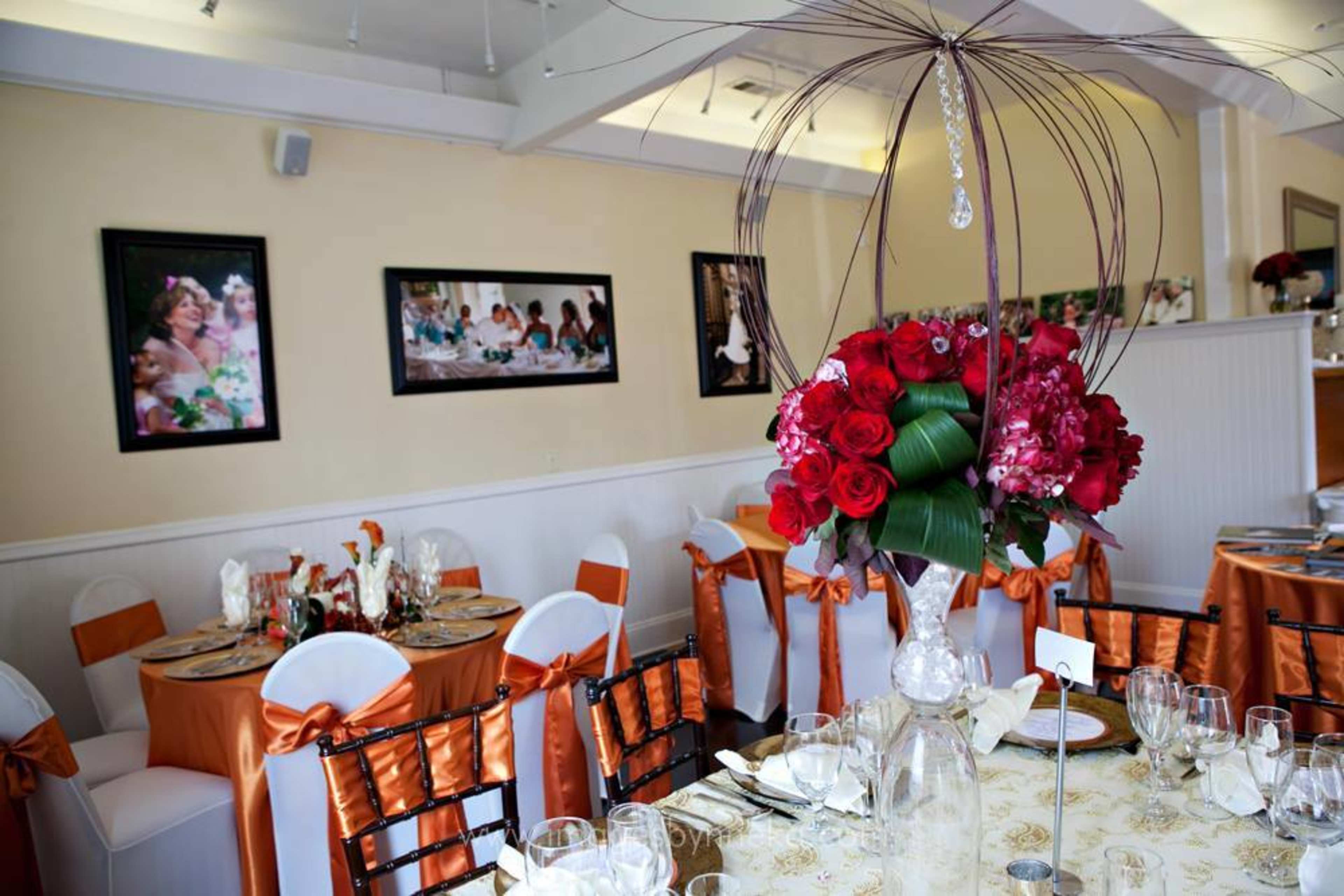 An elegantly set dining table with orange and white decorations, featuring floral centerpieces and framed photographs on the walls.