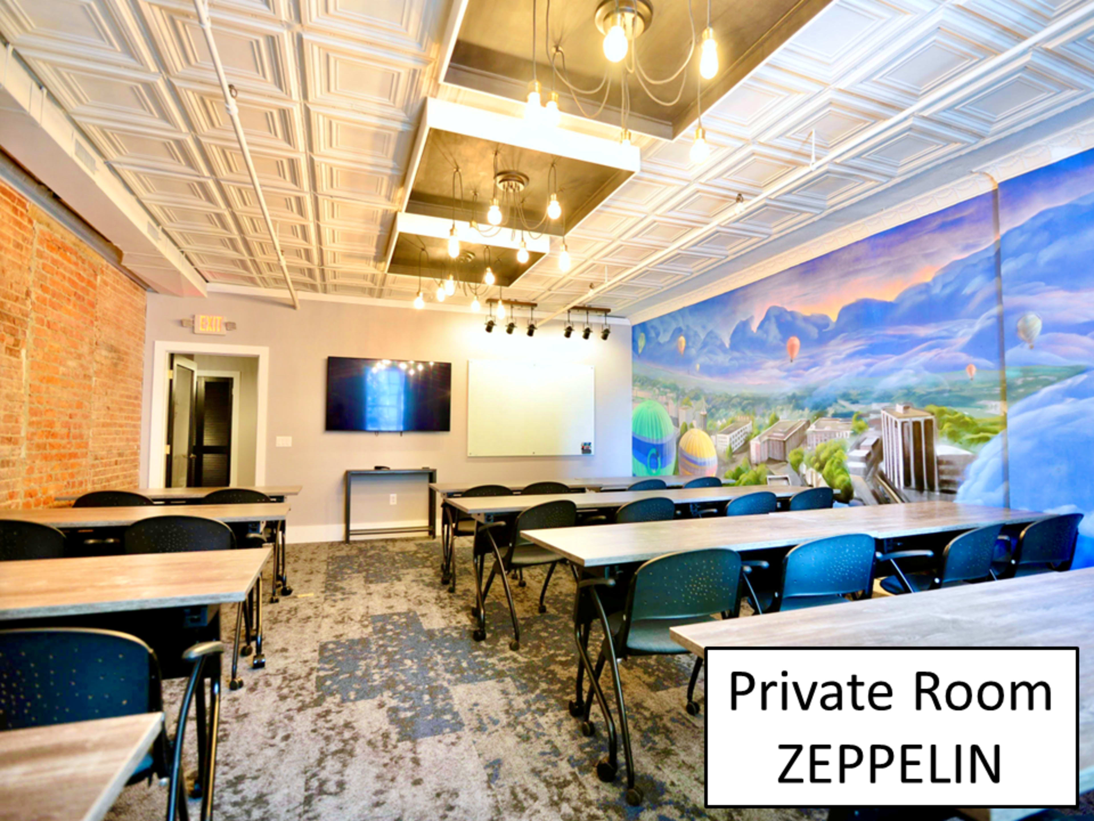 A private meeting room with tables arranged in rows, a large wall mural of mountains and hot air balloons, and a television mounted on the wall.