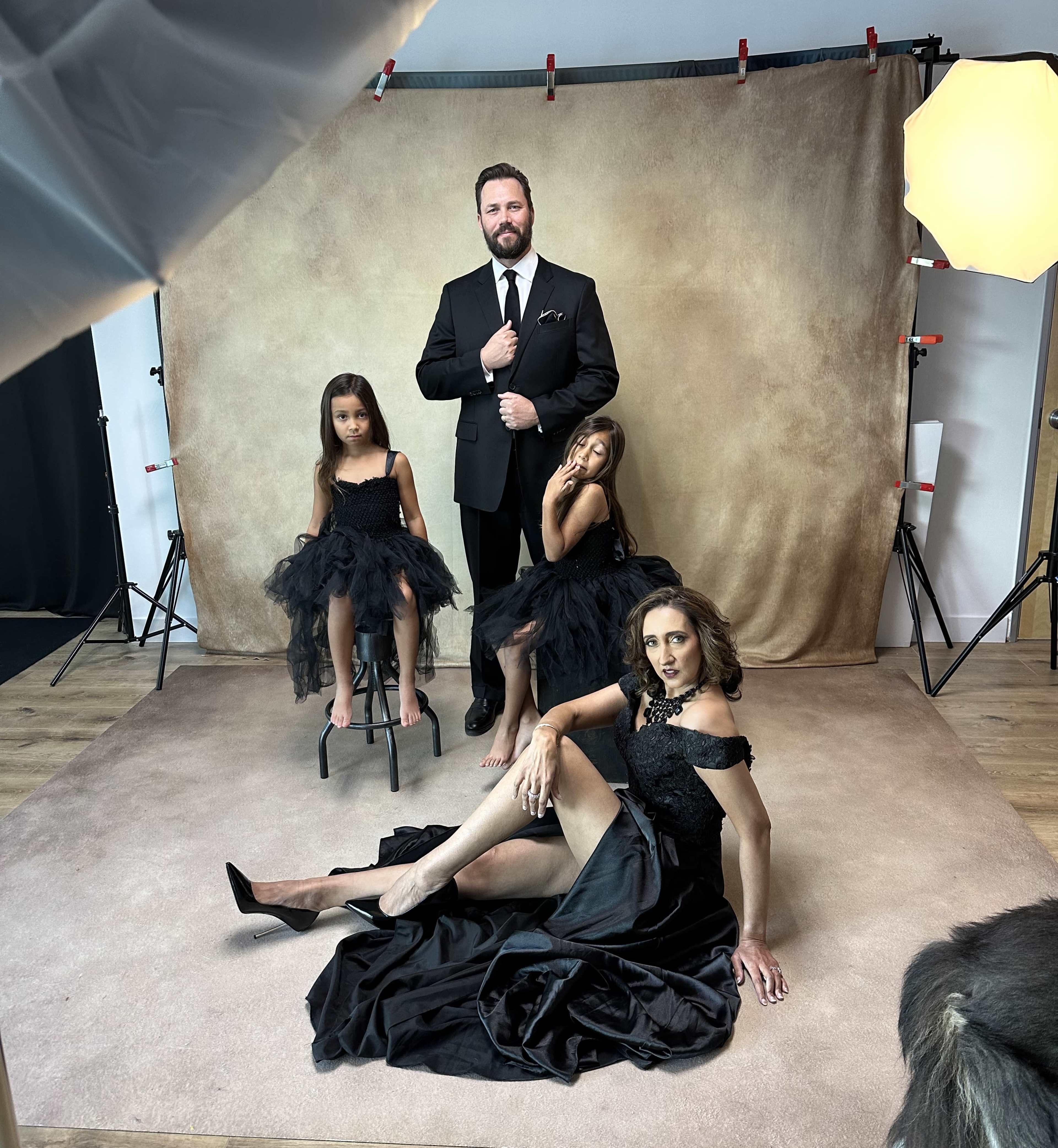 A man in a black suit stands with two girls in black dresses, while a woman in a black gown poses on the floor in front of them, all against a neutral backdrop.