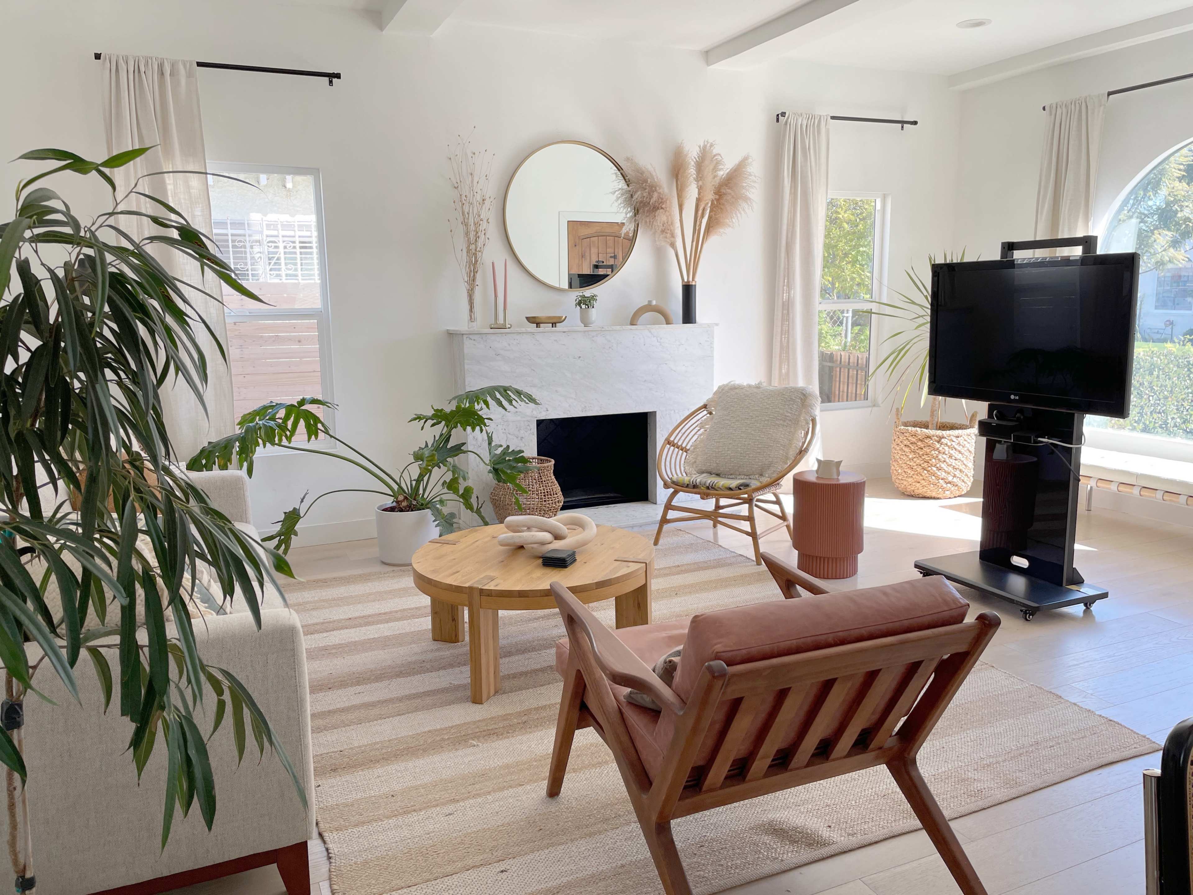 The image shows a bright, modern living room featuring a circular mirror above a white fireplace, a round wooden coffee table, a plant, and various seating options.