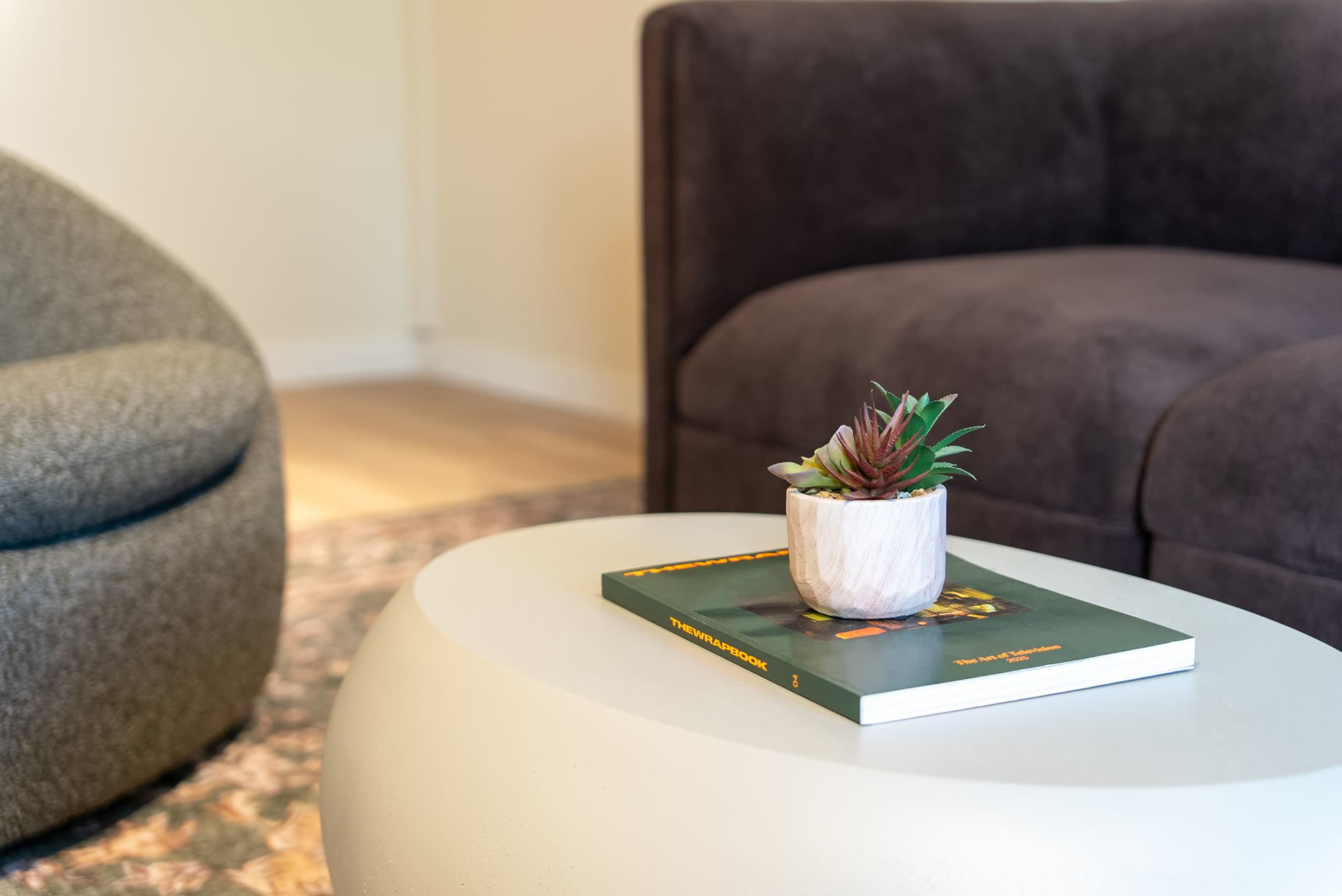 A small plant in a decorative pot sits atop a book on a round coffee table, next to a gray upholstered chair and a dark couch.