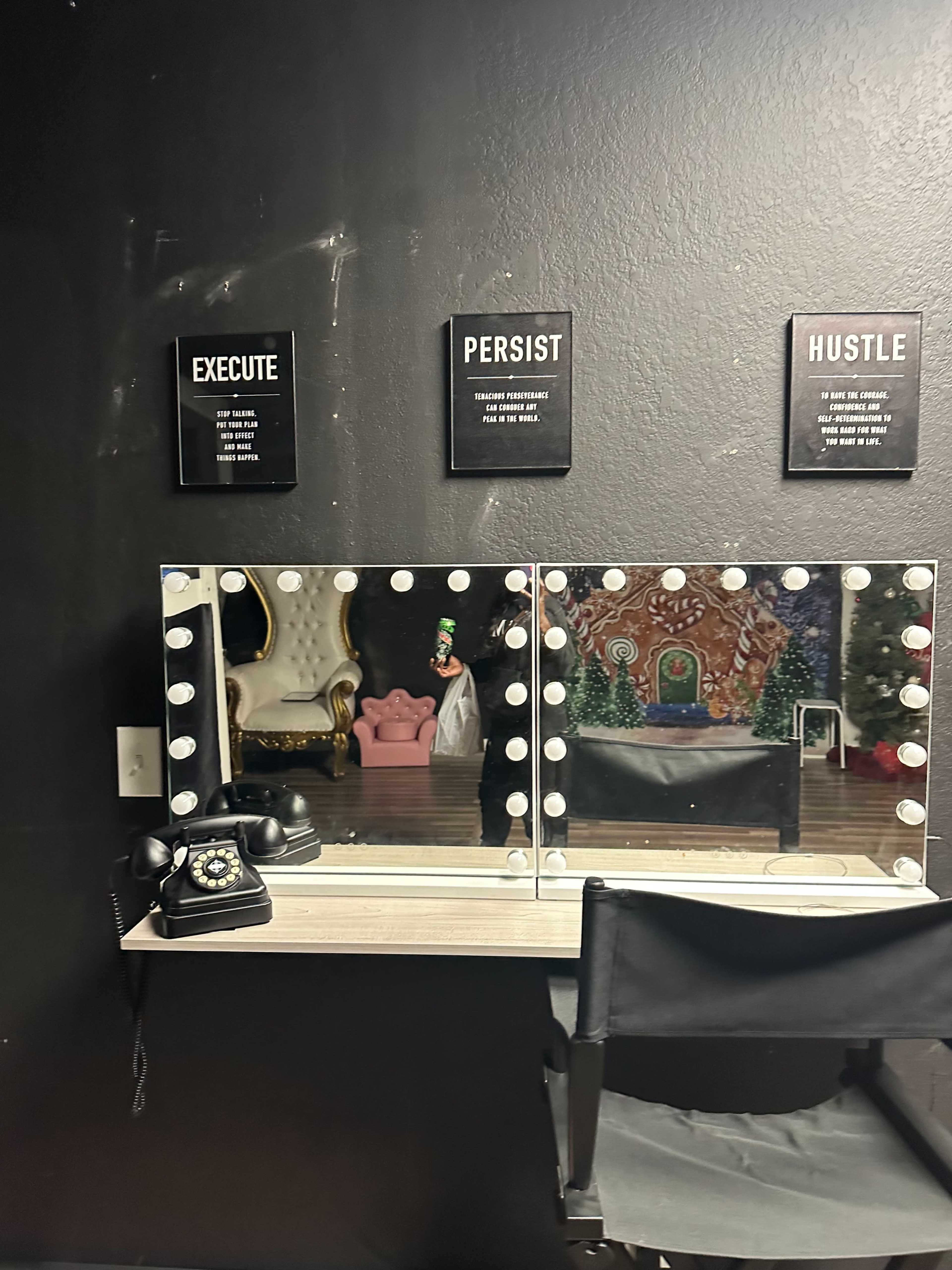The image shows a vanity mirror with bright lights, a black chair, a vintage telephone, and motivational signs on the wall.