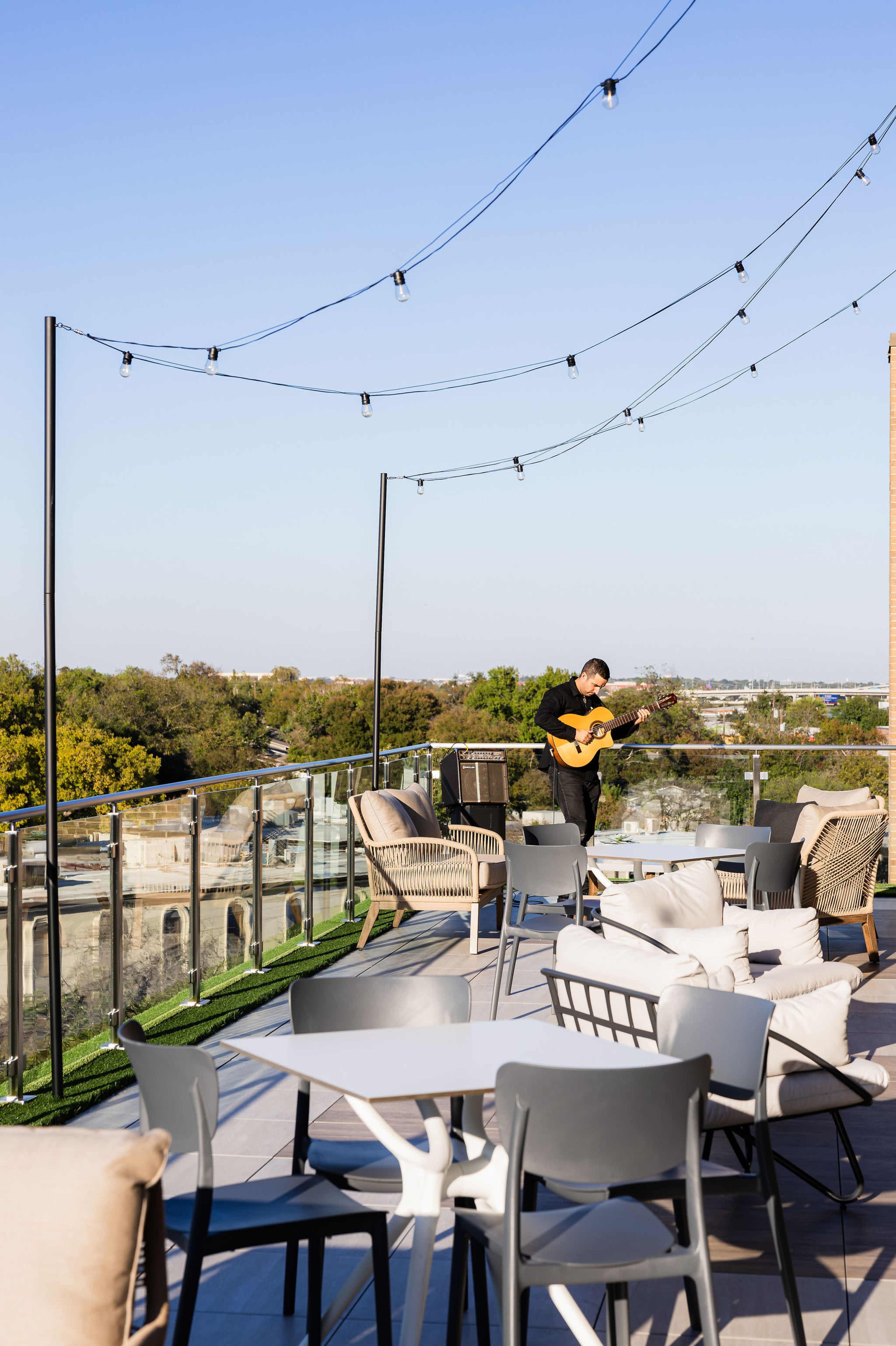 A musician plays an acoustic guitar on a rooftop terrace adorned with string lights and modern seating arrangements.