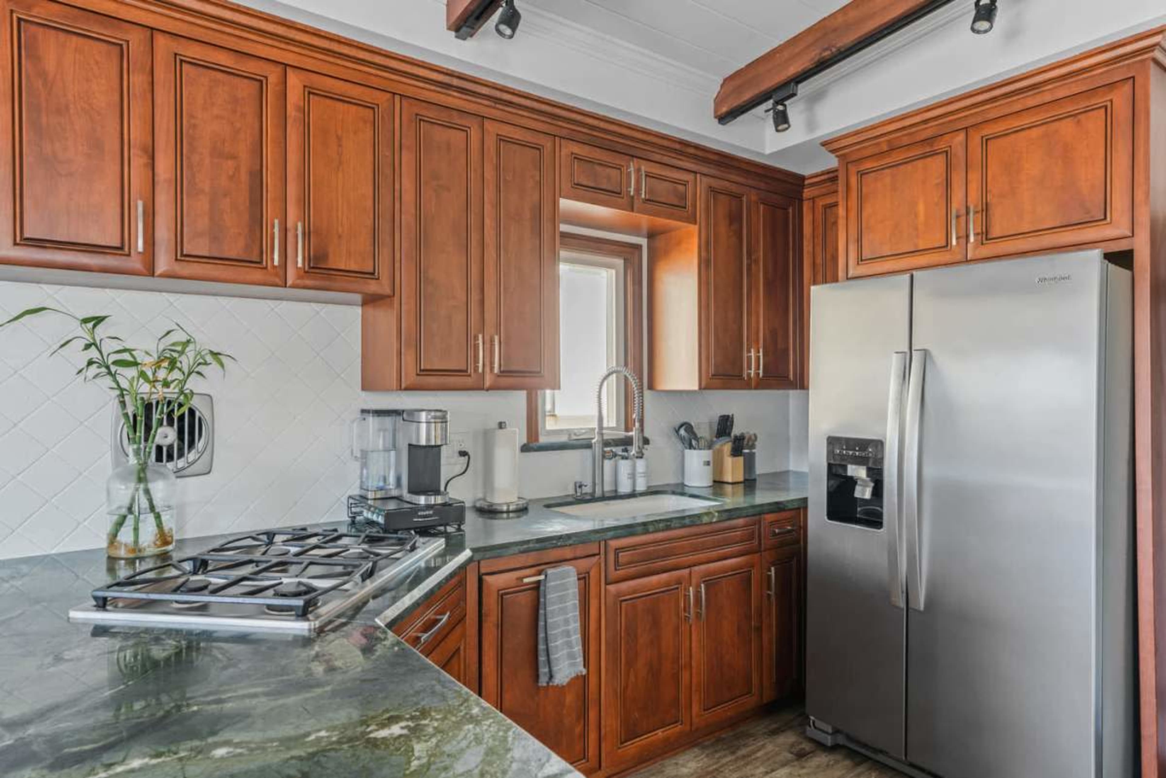 The image shows a modern kitchen with wooden cabinets, a stainless steel refrigerator, and a countertop with a gas stove.