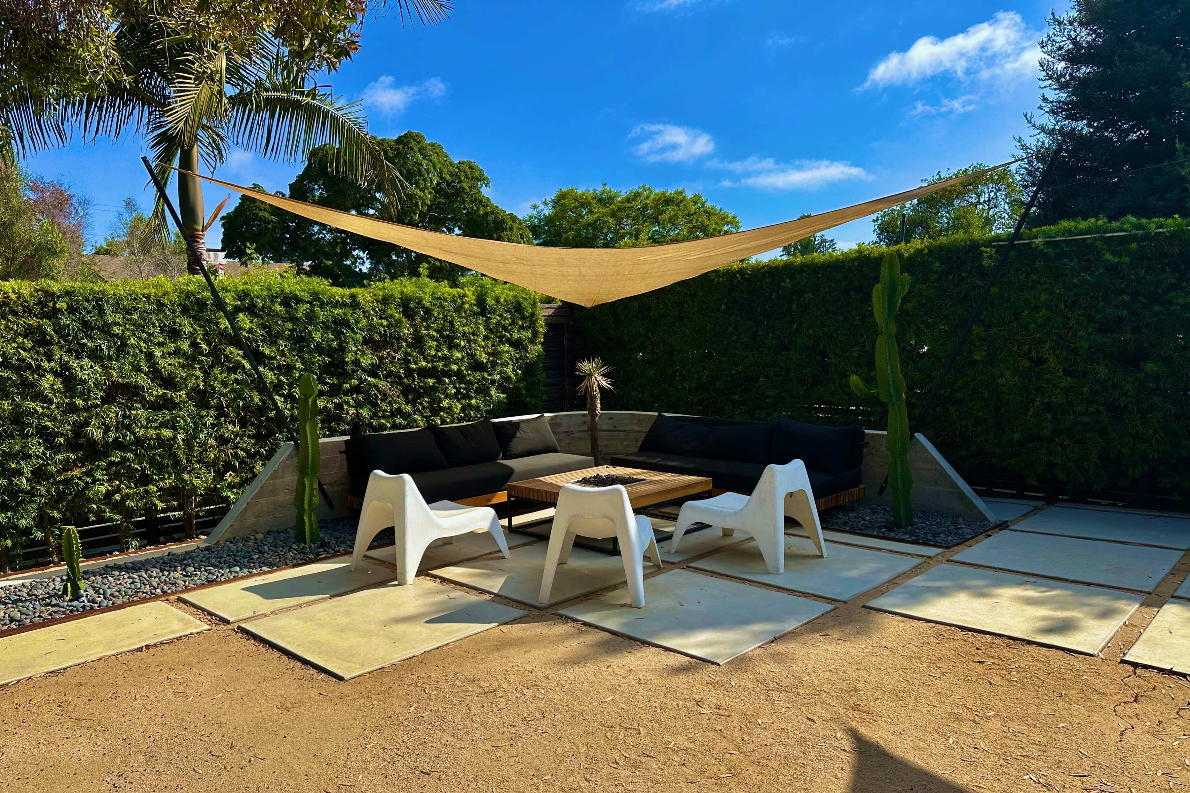 A shaded outdoor seating area features black couches around a wooden table on a stone patio, surrounded by greenery and cacti.