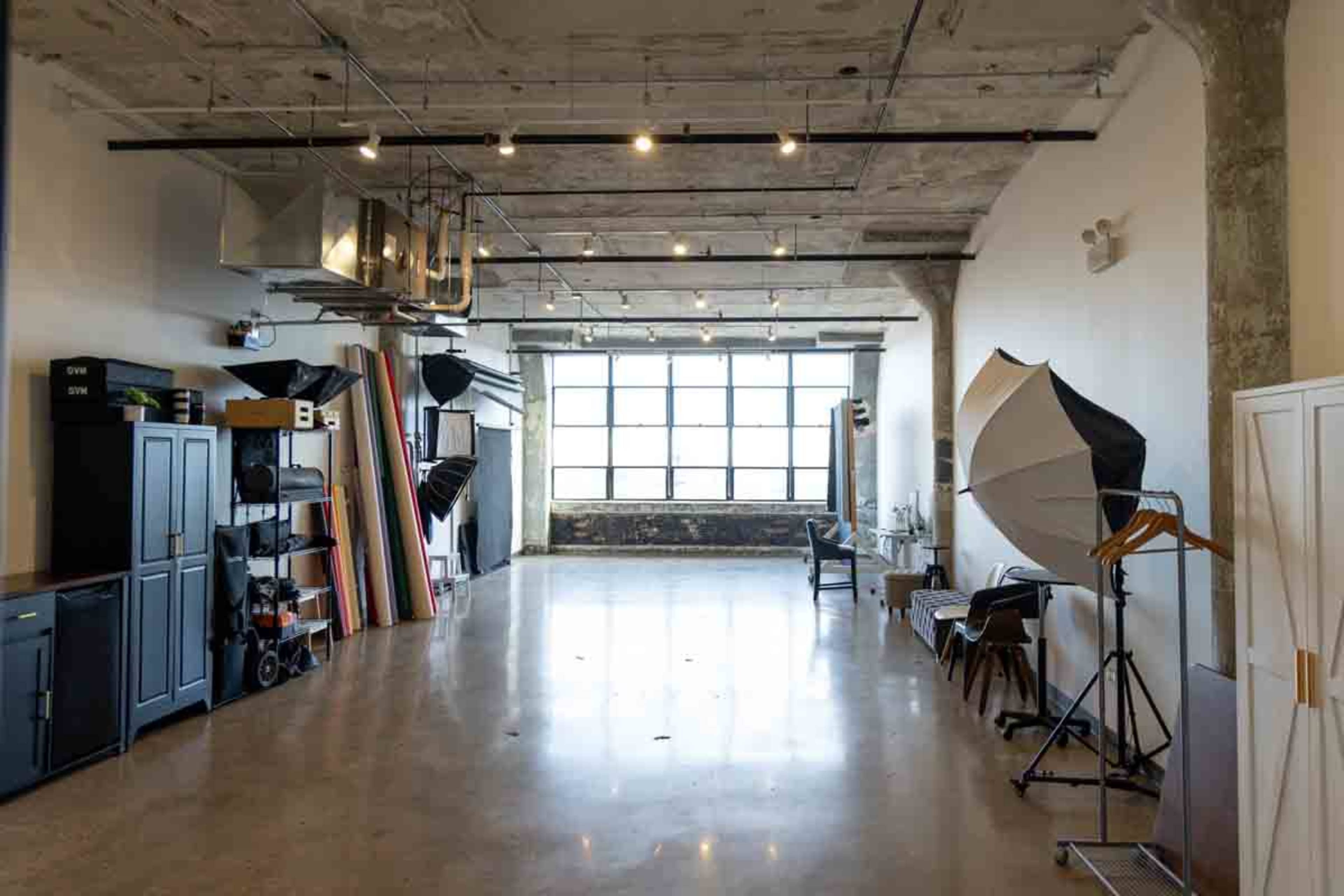 The image depicts a spacious, industrial-style studio with large windows, various photography equipment, and minimalist furnishings.