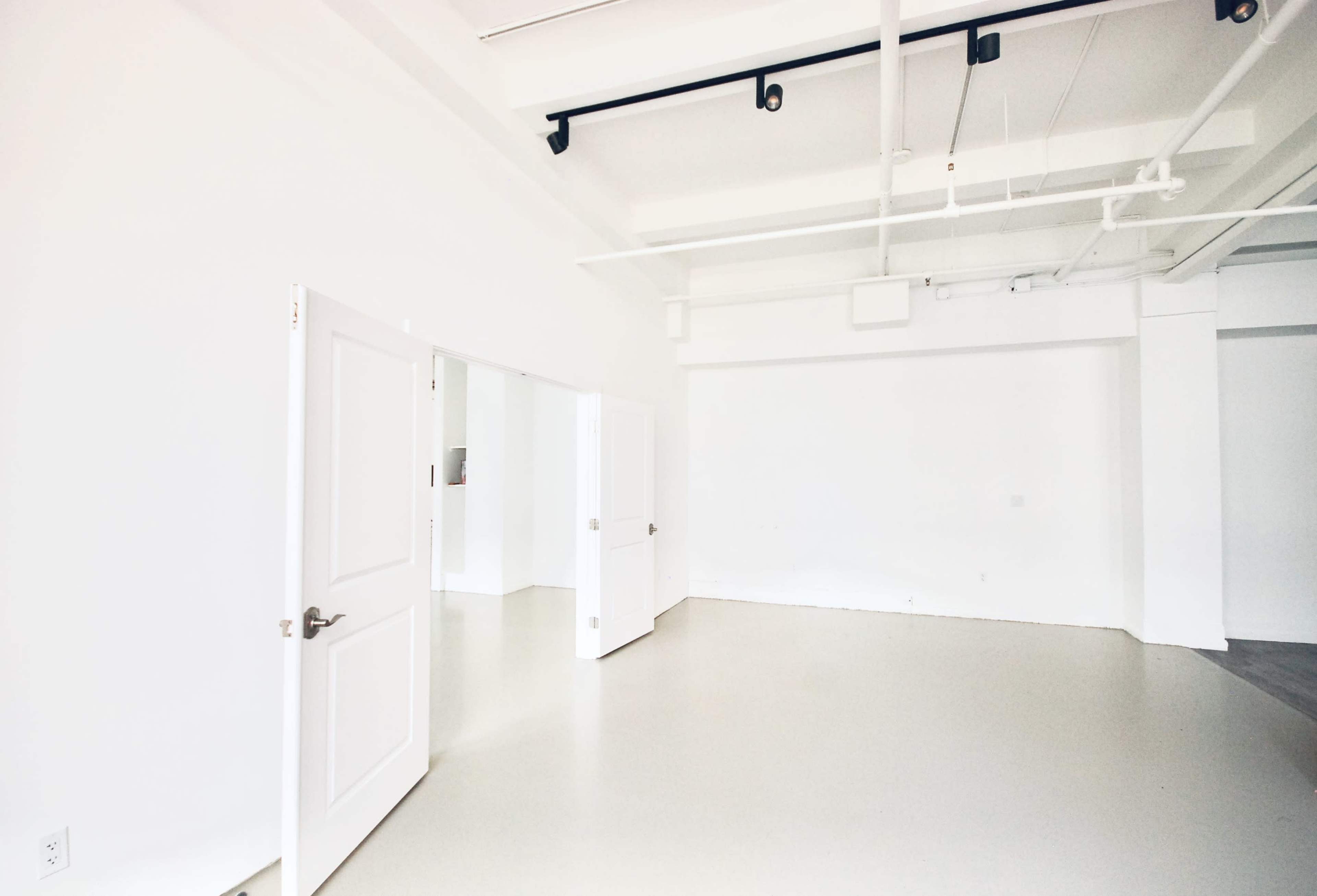 An empty room with white walls, light flooring, and two open doors leading to adjacent spaces.