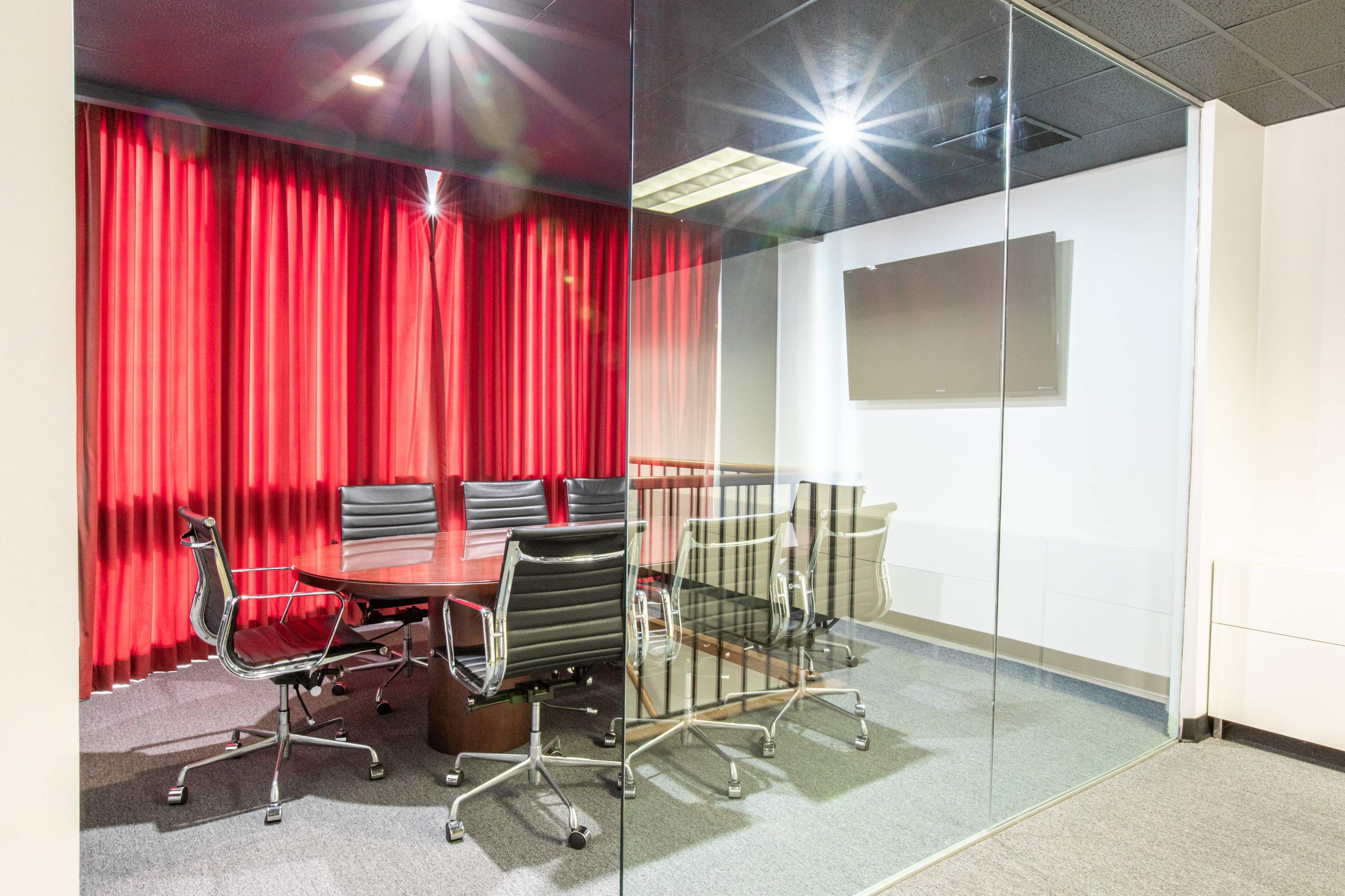 A glass-walled conference room features a large round table surrounded by black chairs and is illuminated by red curtains draping in the background.
