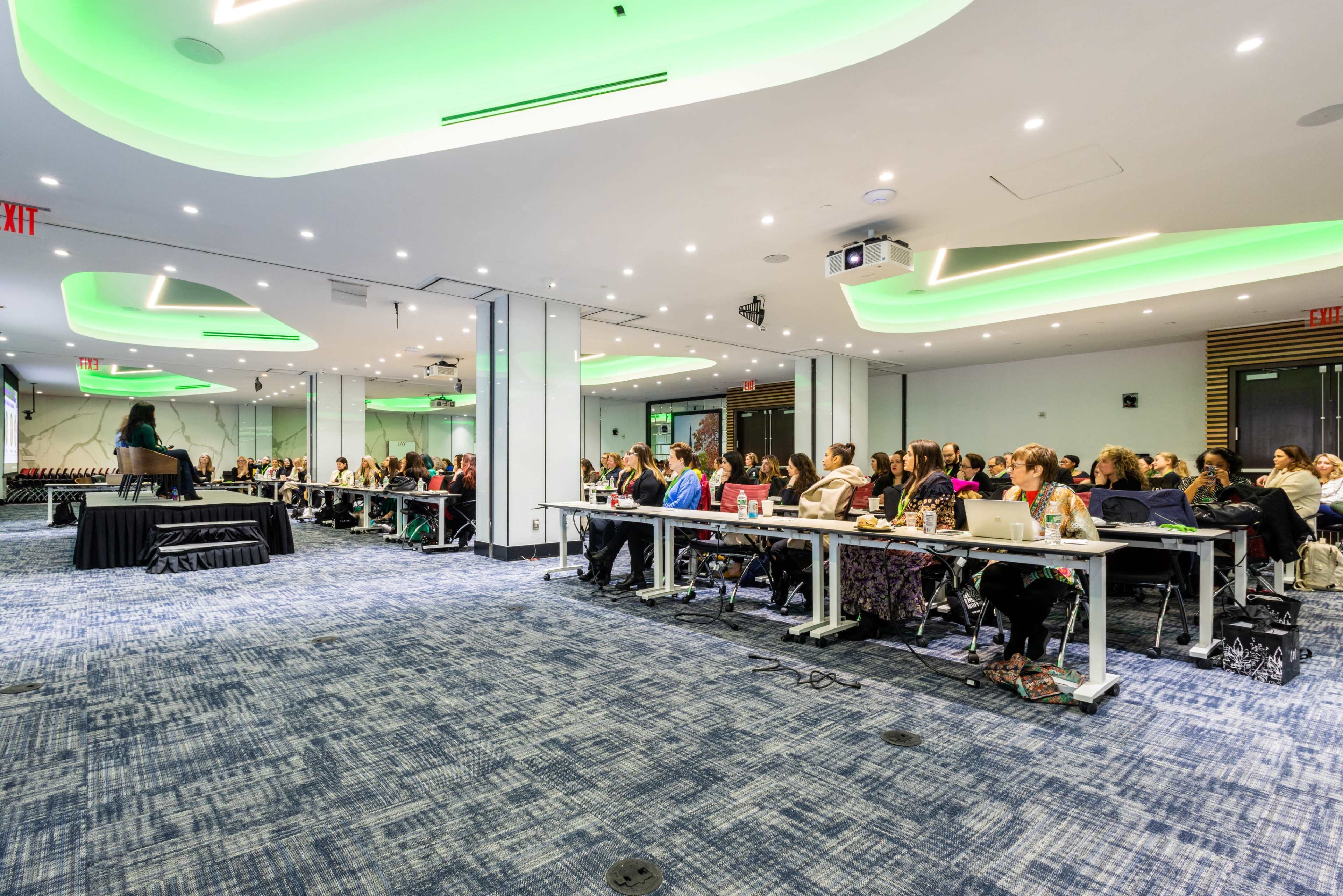 A conference room setup features rows of tables with attendees seated and focused on a speaker at the front.