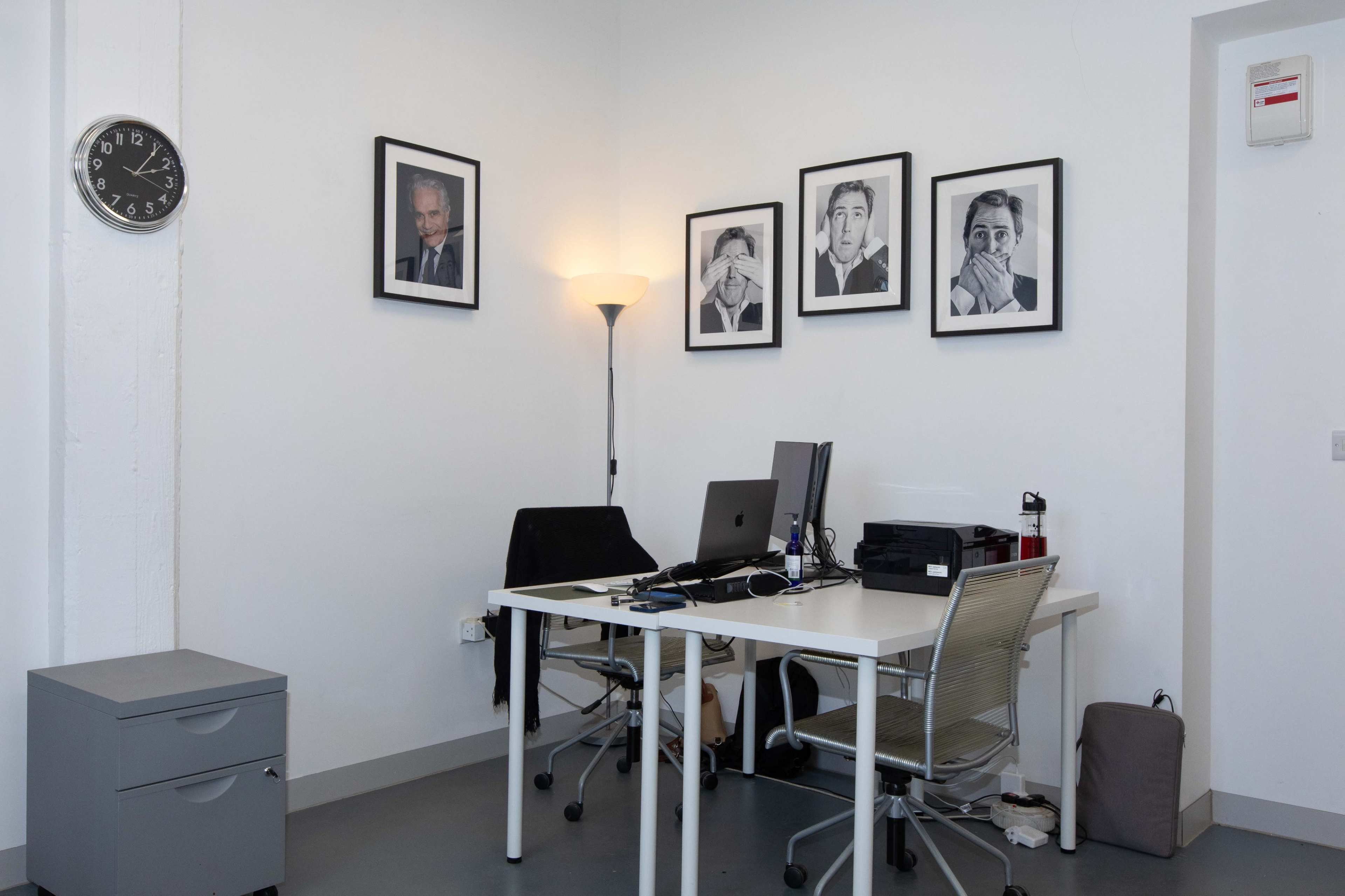 A neatly arranged office space features a desk with a computer, a chair, and four framed portraits on the wall, along with a clock and a filing cabinet.