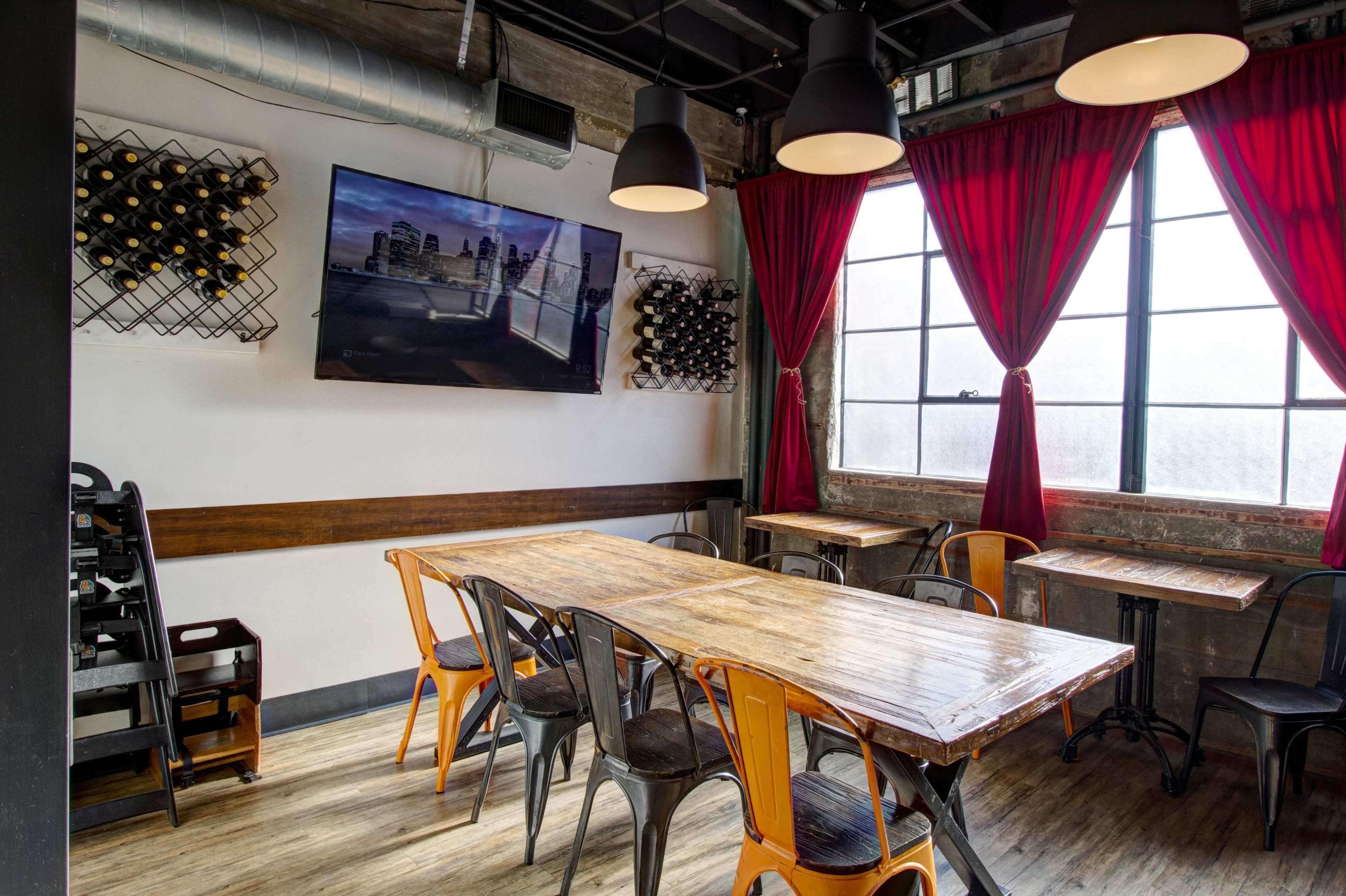 A dining area features a wooden table surrounded by metal chairs, with a large television mounted on the wall and red curtains framing a window.