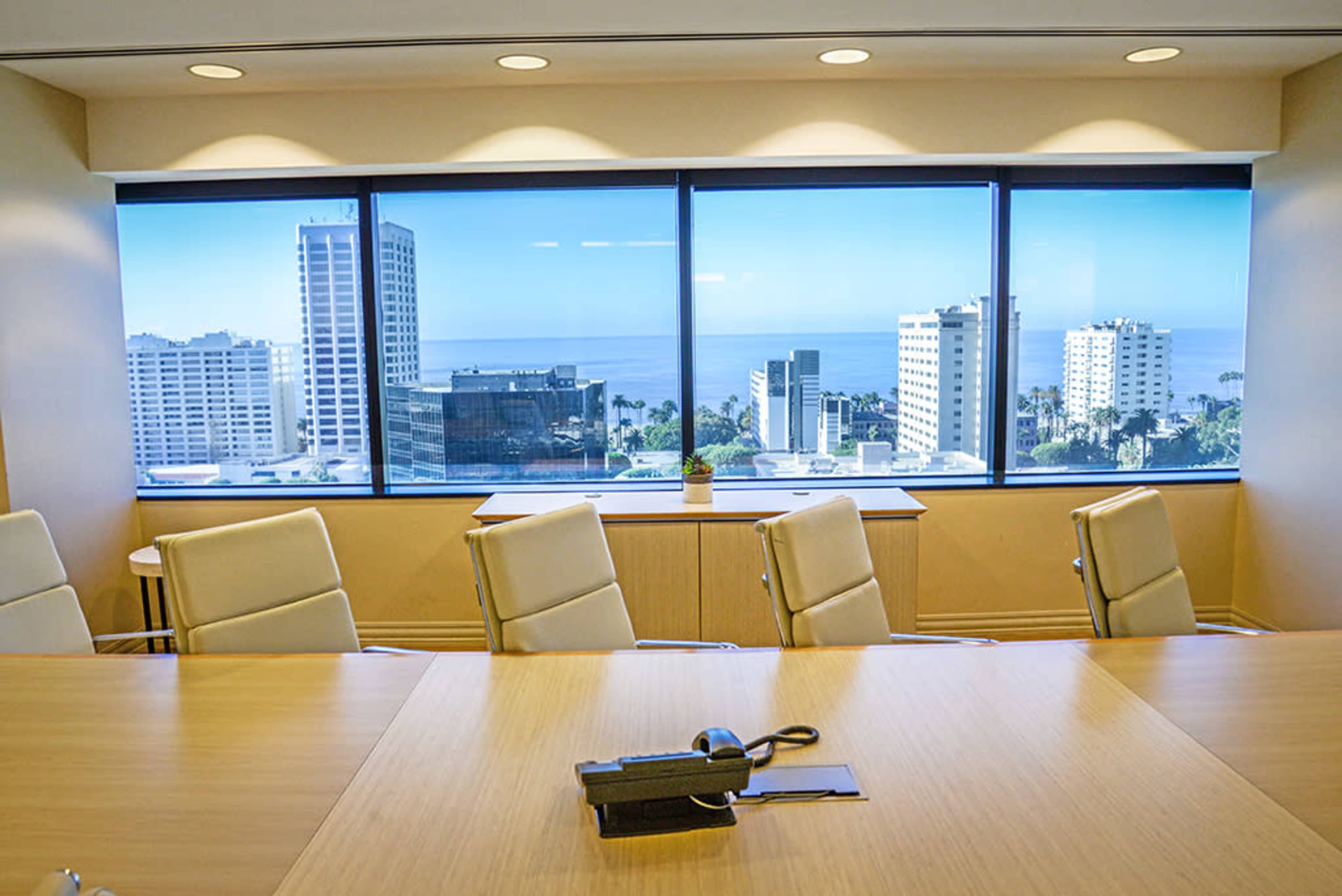 A conference room with a wooden table, white chairs, and large windows overlooking a coastal city skyline.