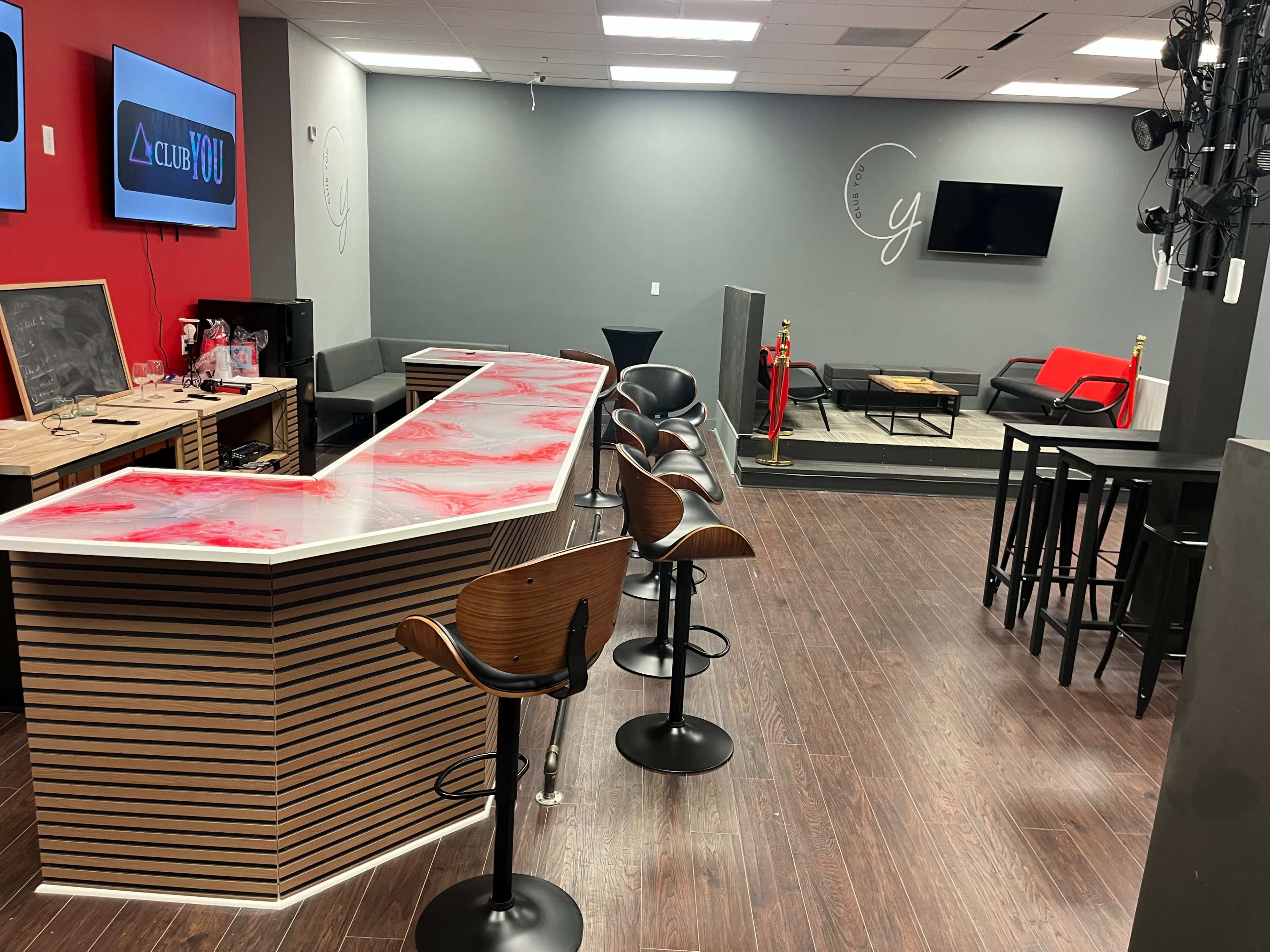 The image shows a modern lounge area with a bar, high tables, and seating arrangements, featuring a mix of red and neutral-colored walls.