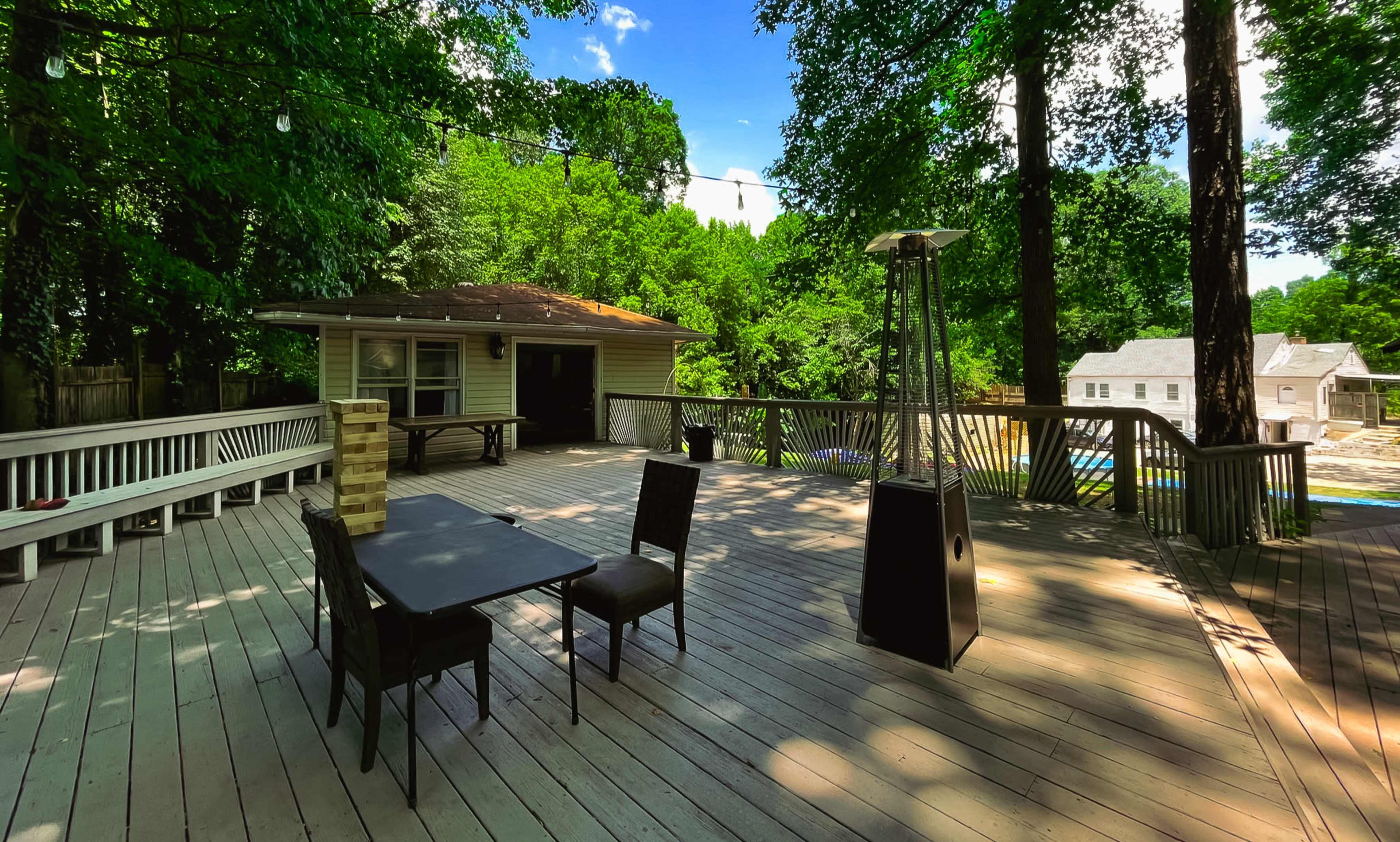 A spacious wooden deck features a table and chairs, surrounded by trees and overlooking a nearby pool and buildings.