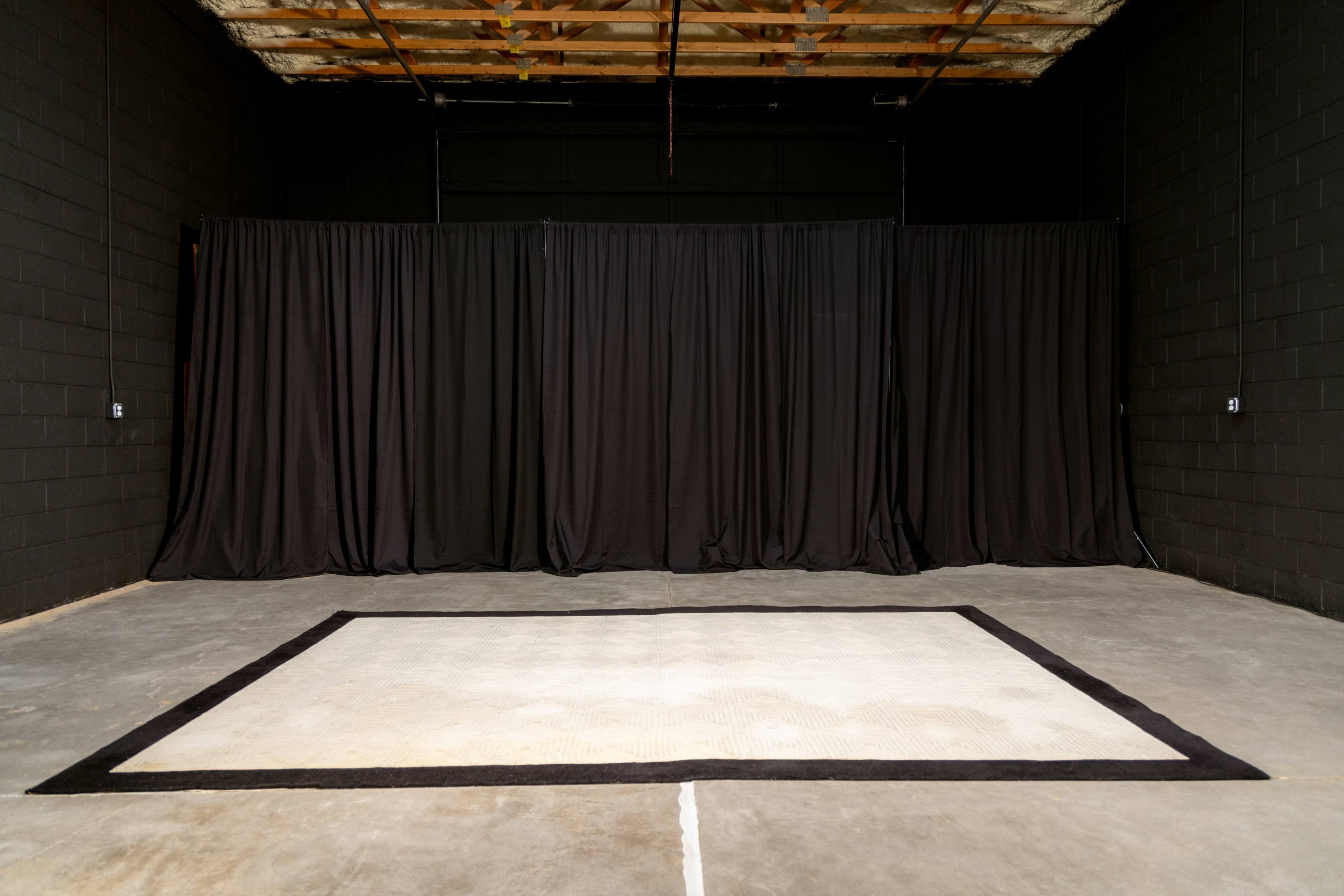 An empty room features a large, bordered rug on a concrete floor, with black curtains draping the walls.