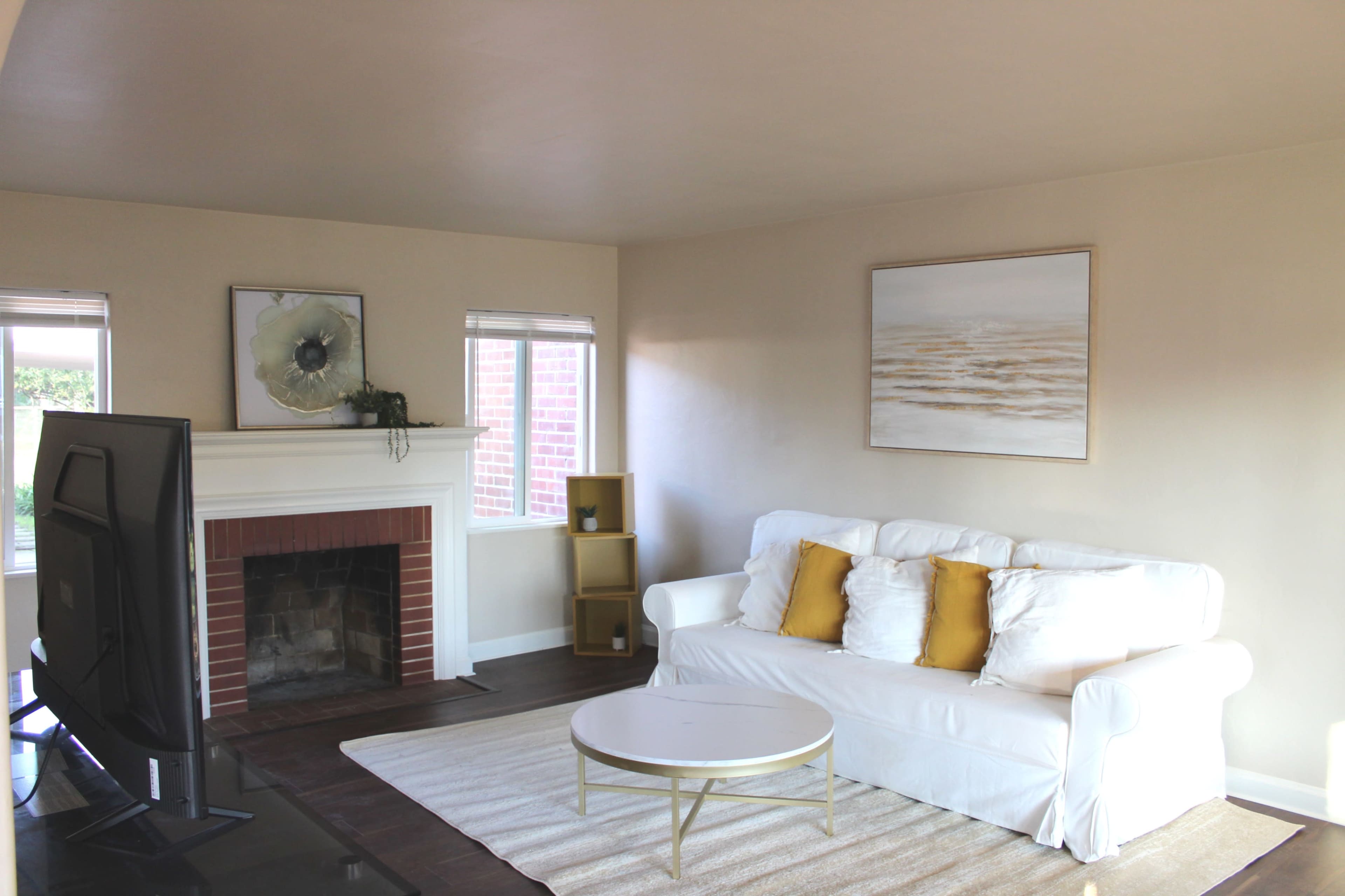 A light-filled living room featuring a white sofa with yellow cushions, a round coffee table, a fireplace, and a large window.