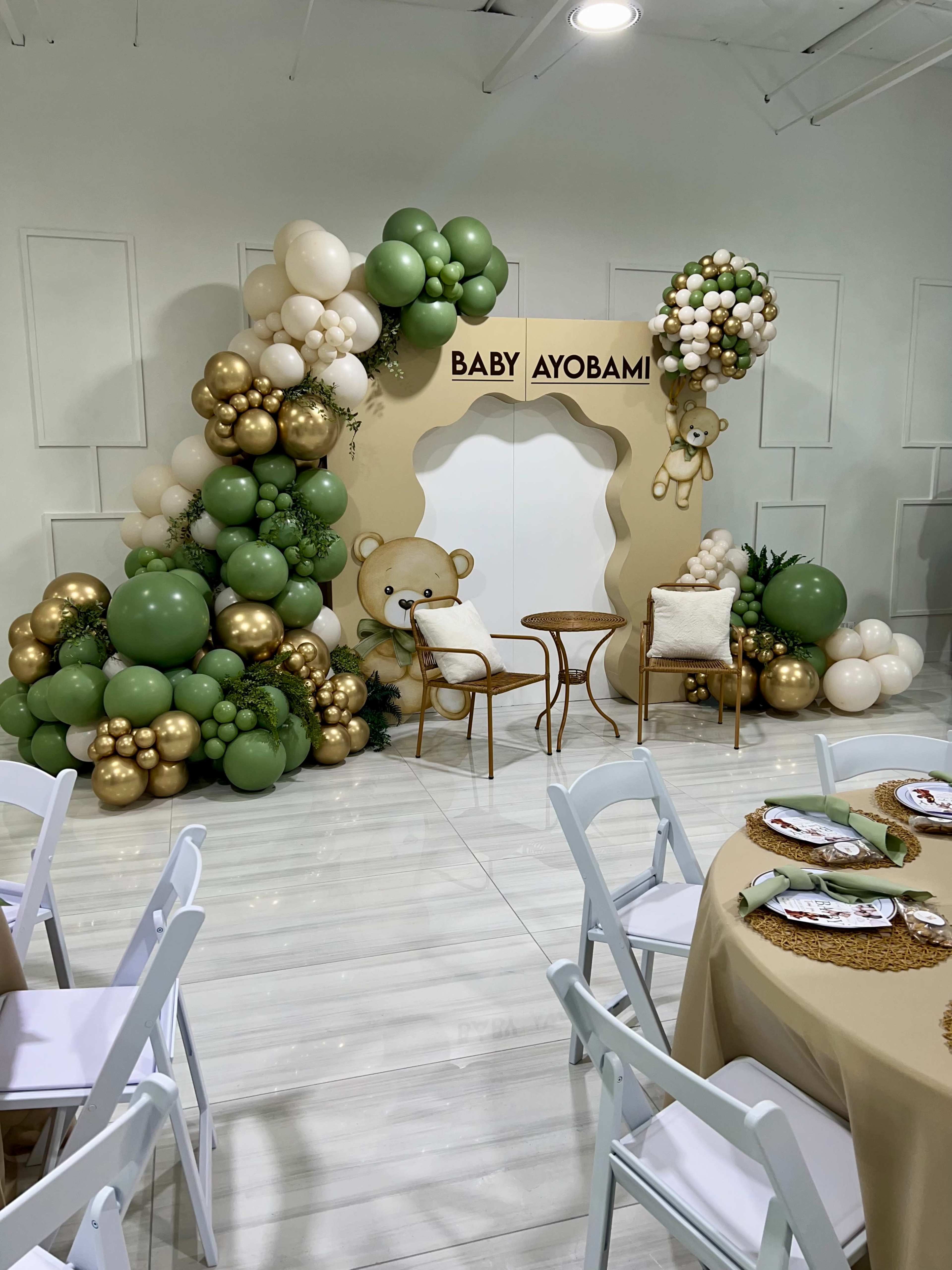 A decorative backdrop featuring balloons in various colors, a bear illustration, and the words "BABY AYOBAMI," set up for a celebration, with chairs and tables arranged in the foreground.