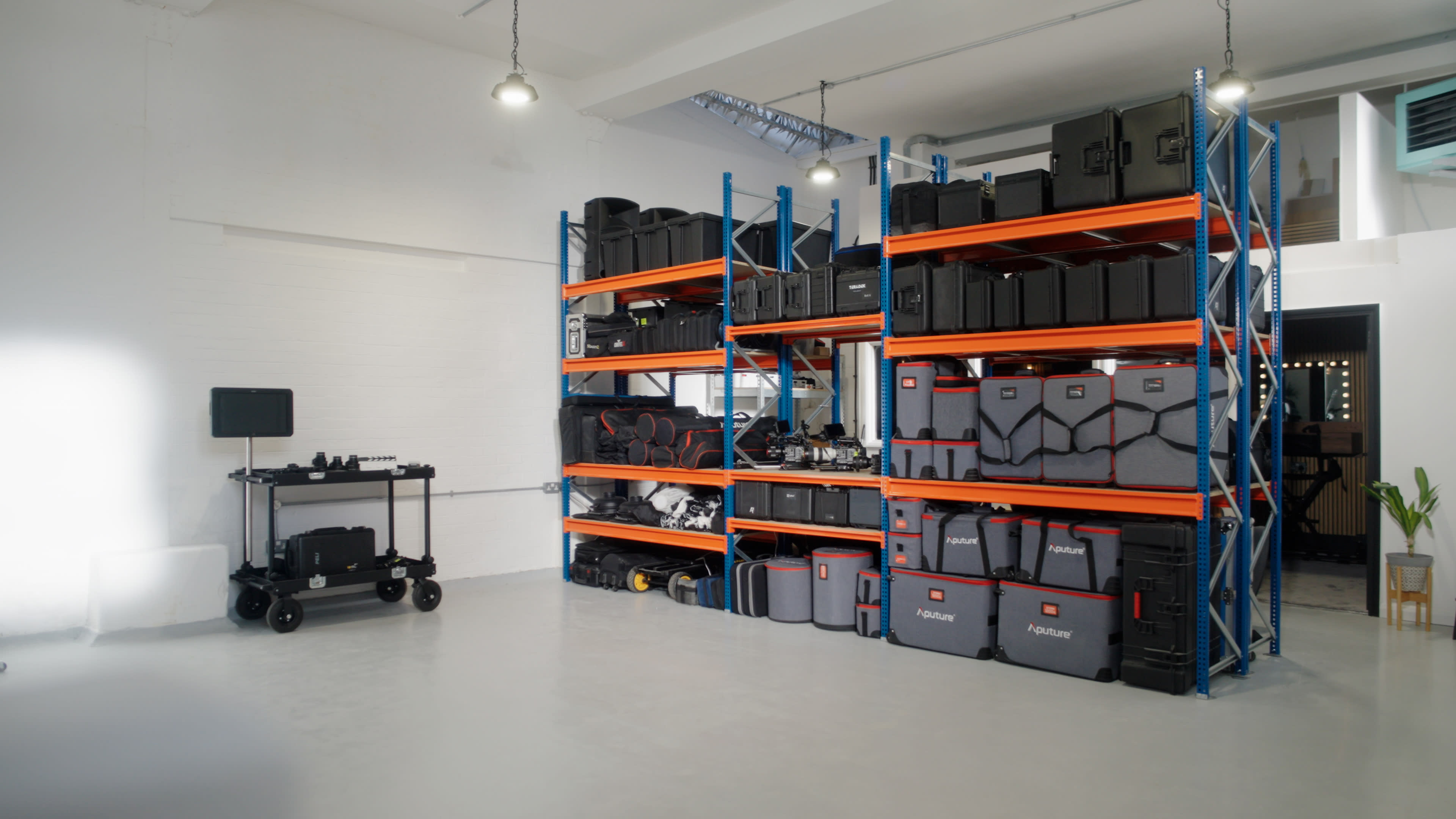 The image shows a storage area with metal shelves filled with various bags and cases organized neatly, alongside a small black cart on wheels.