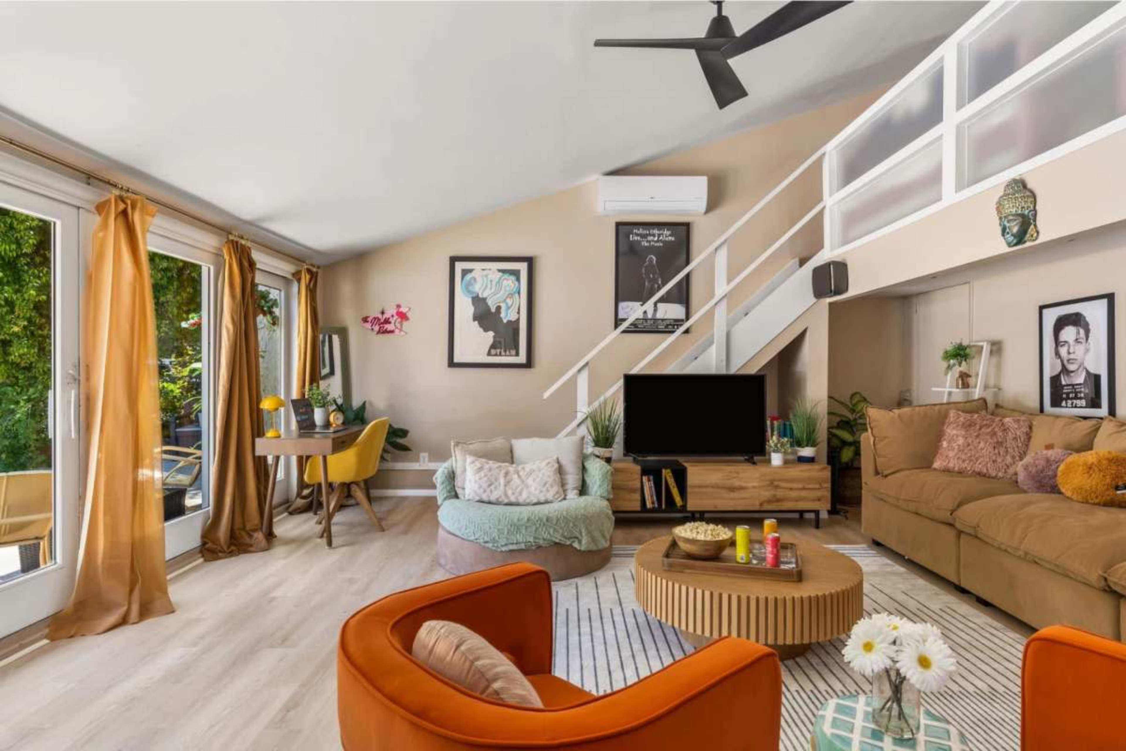 The image shows a modern living room with a beige color scheme, featuring large windows, a comfortable couch, a circular coffee table, and a dining area with a table and chairs.