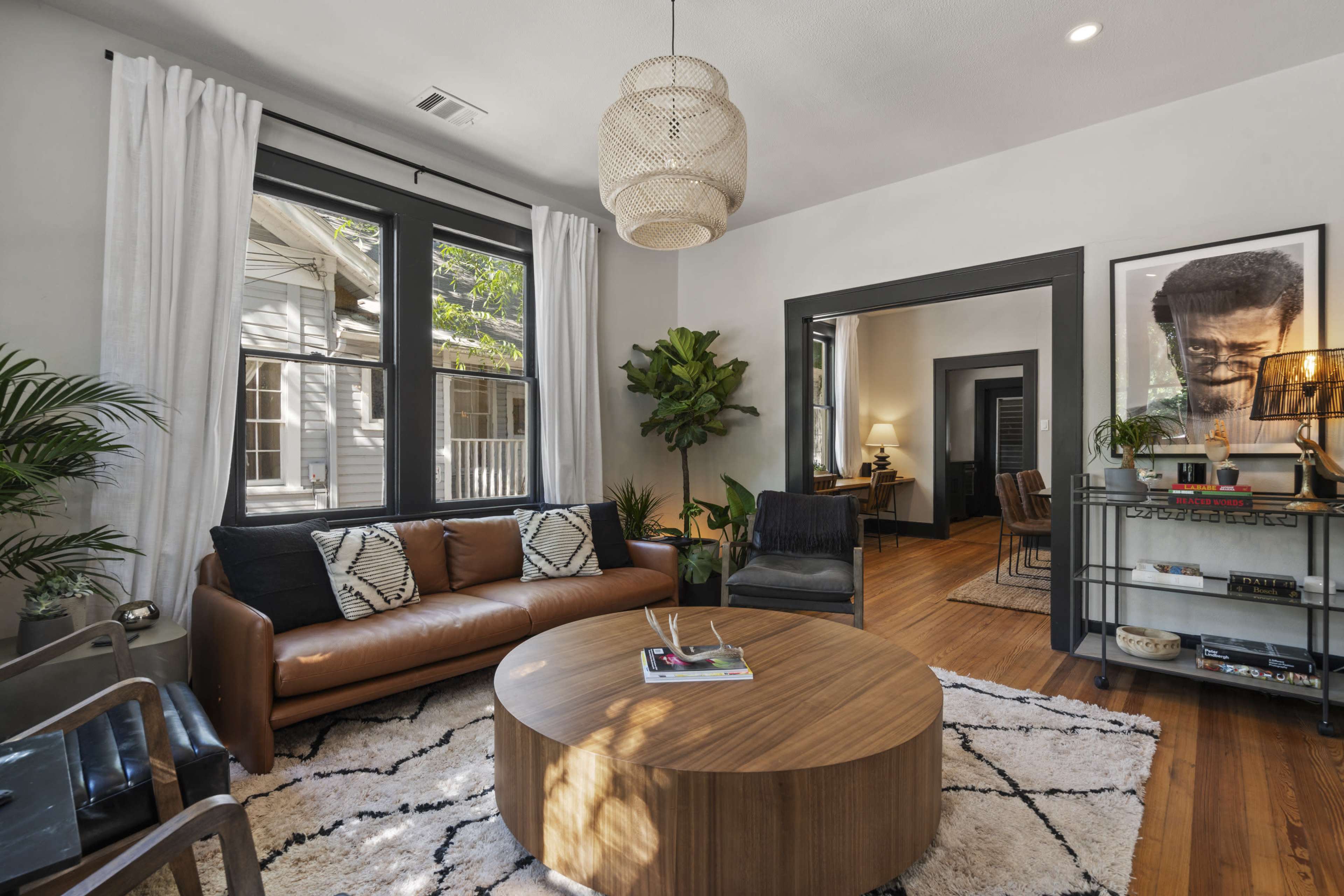 The living room features a leather sofa, a round wooden coffee table, and large windows with white curtains, surrounded by indoor plants and contemporary decor.