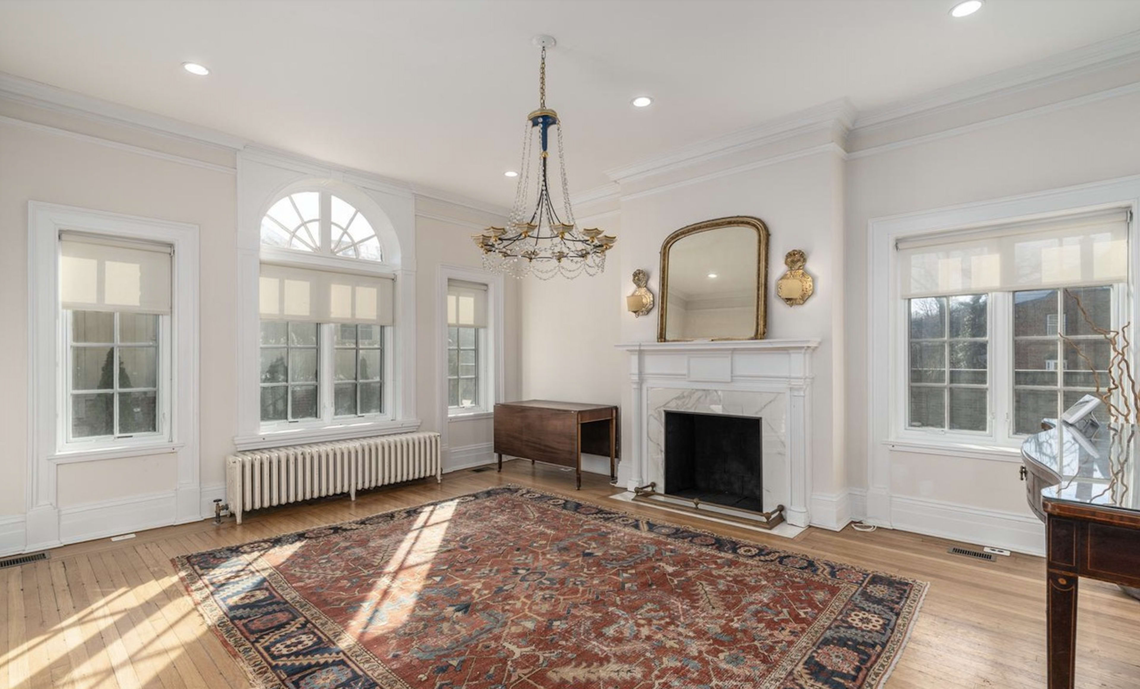The room features a chandelier, large windows, a fireplace, and a patterned area rug on hardwood flooring.