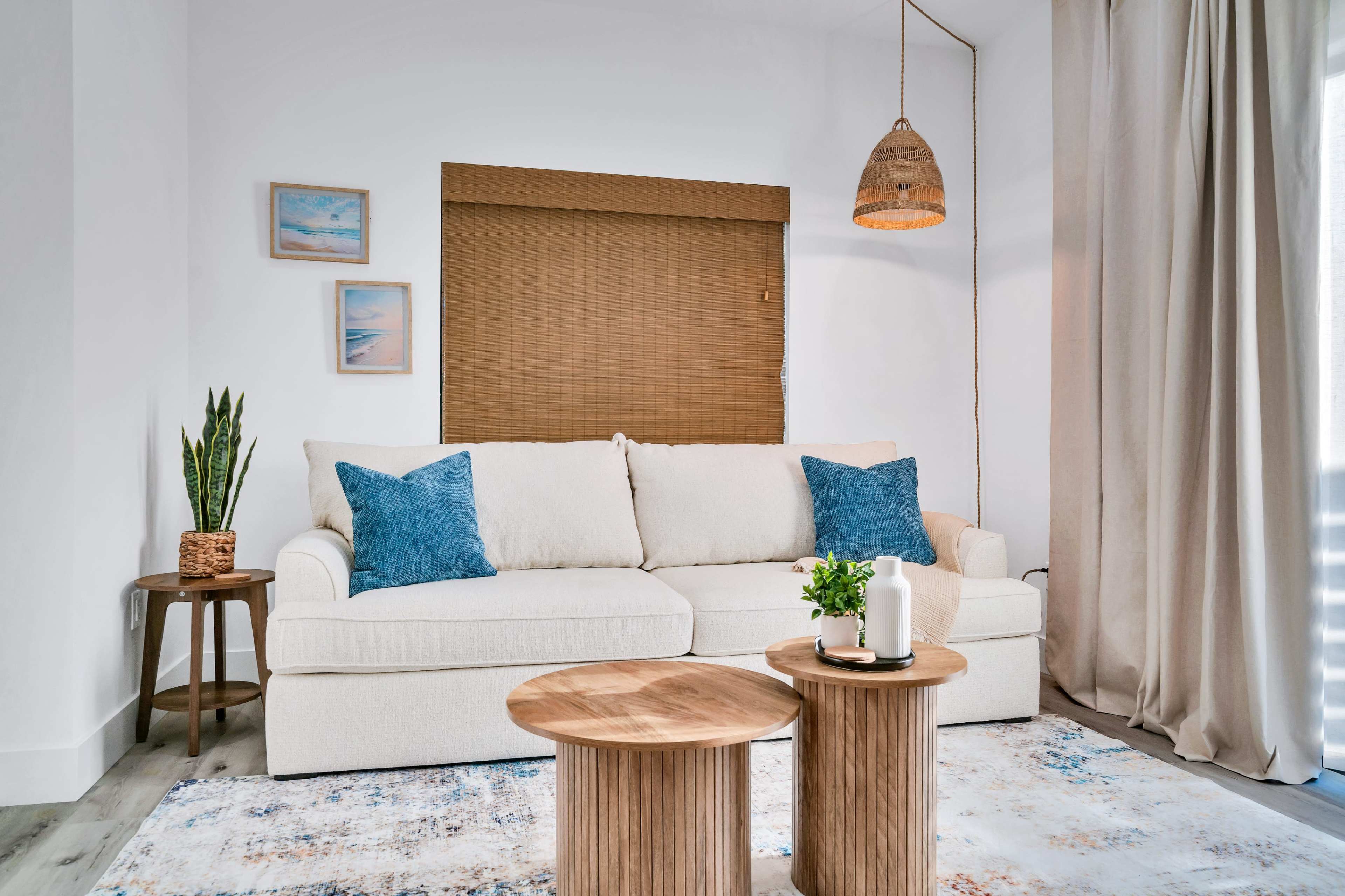 A light-colored sofa with blue cushions sits in front of a window adorned with blinds, next to two wooden side tables and a plant in a woven basket.