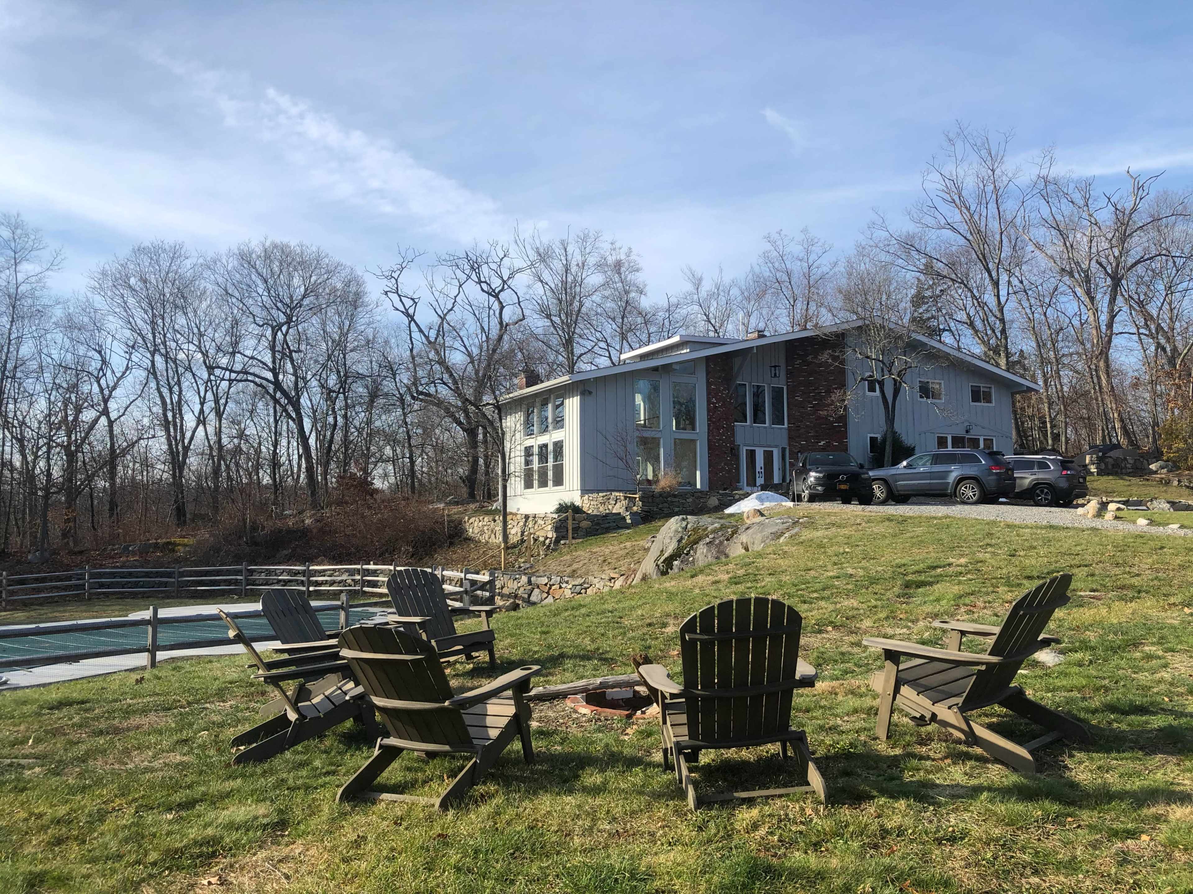 The image shows a modern house with large windows situated on a grassy area, surrounded by bare trees and a fire pit with wooden chairs in the foreground.