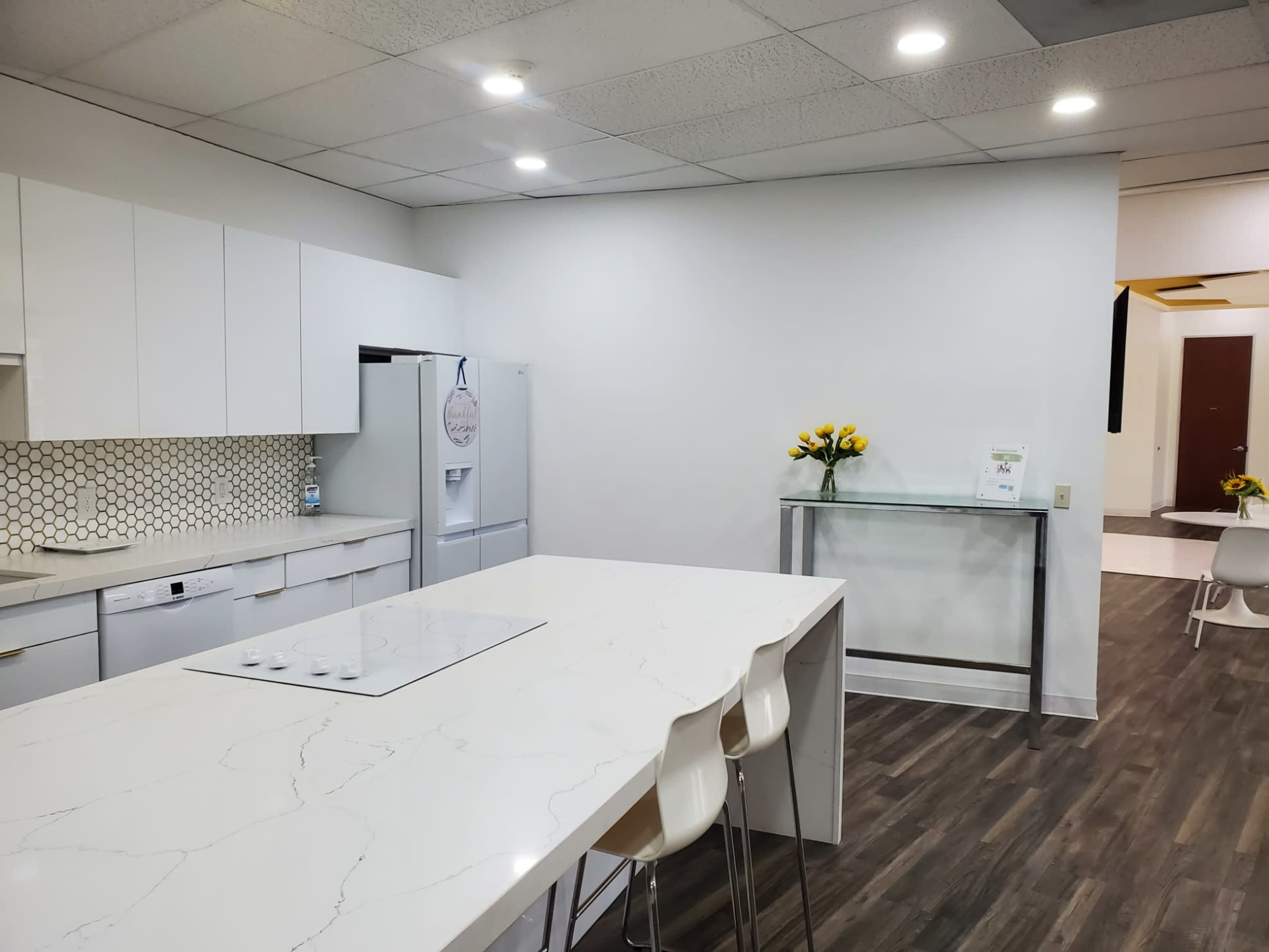 A modern kitchen with a large island, a refrigerator, and a glass table, featuring white cabinetry and a hexagonal tile backsplash.