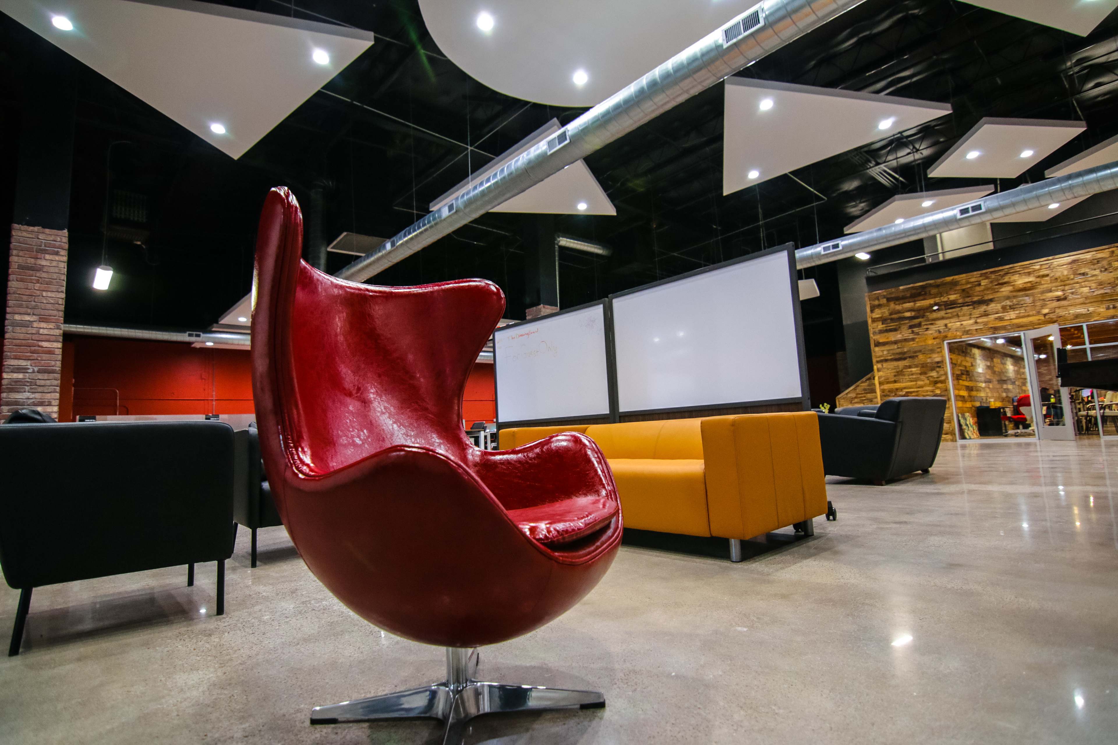 A modern interior space featuring a bright red chair, two couches, and large whiteboards on a polished concrete floor.