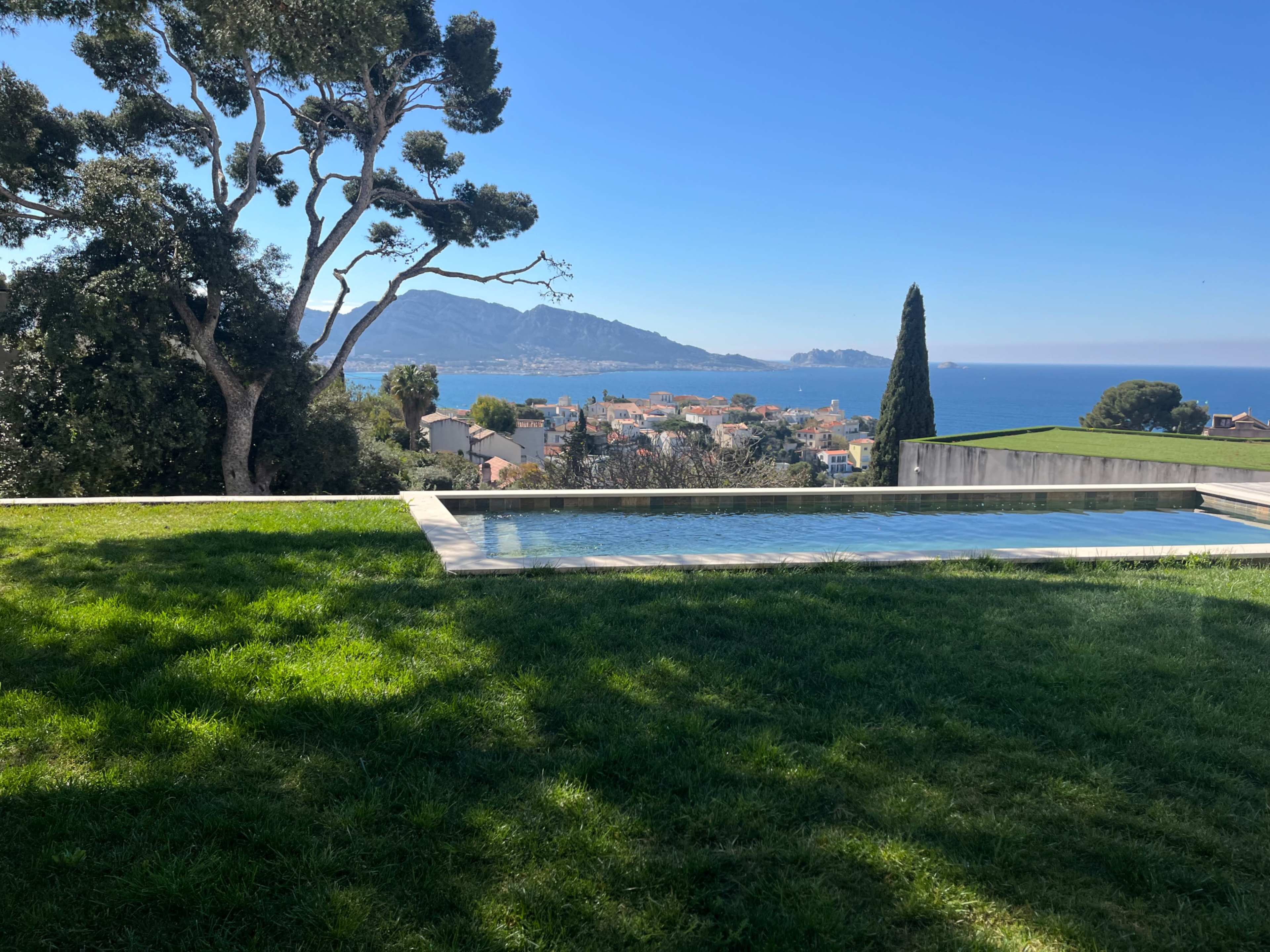 The image shows a view of a pool area with a grassy lawn overlooking a coastal town and the sea, with mountains in the background.