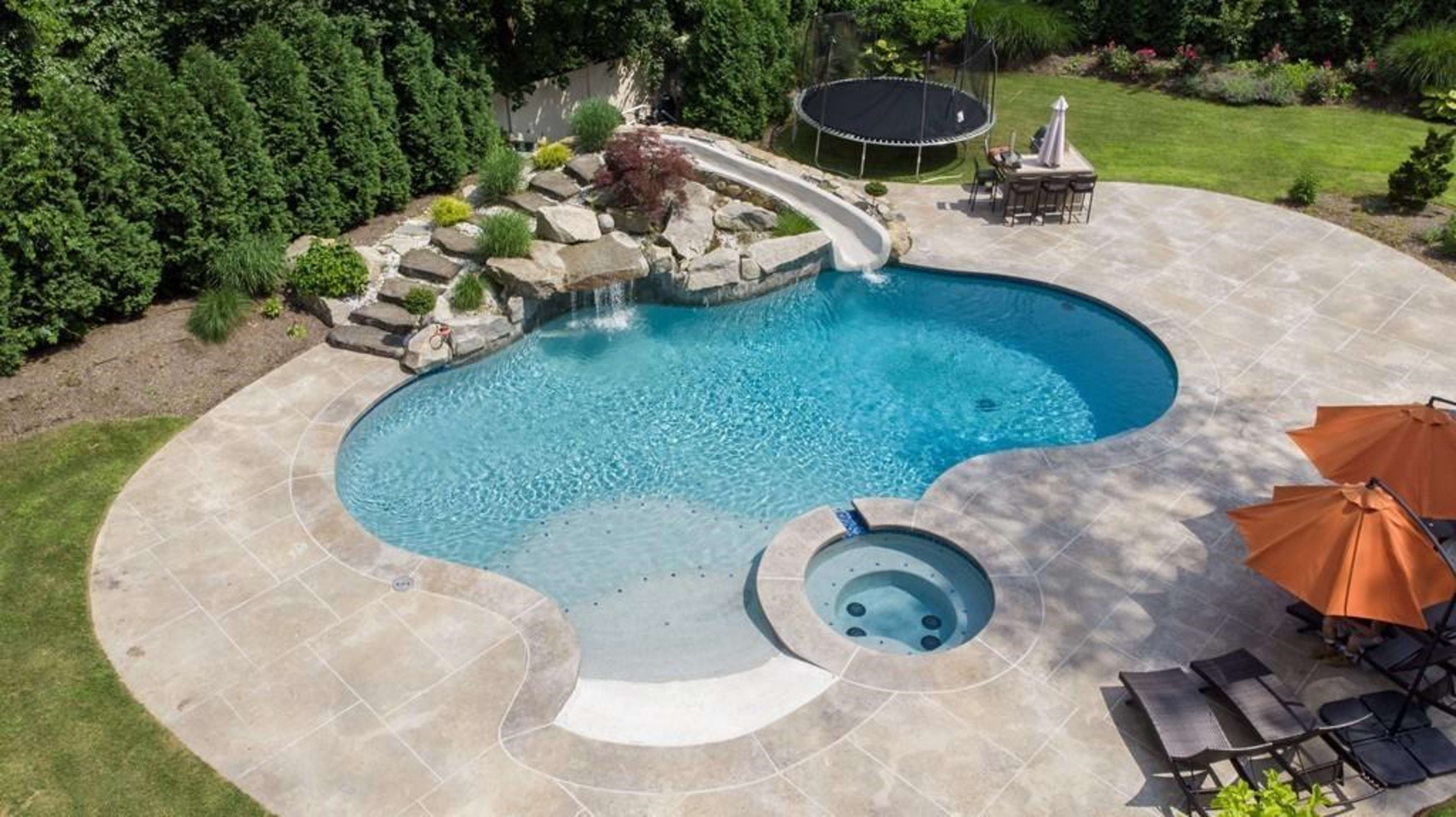 A landscaped backyard featuring a kidney-shaped swimming pool with a slide, a hot tub, a trampoline, and patio furniture under an umbrella.