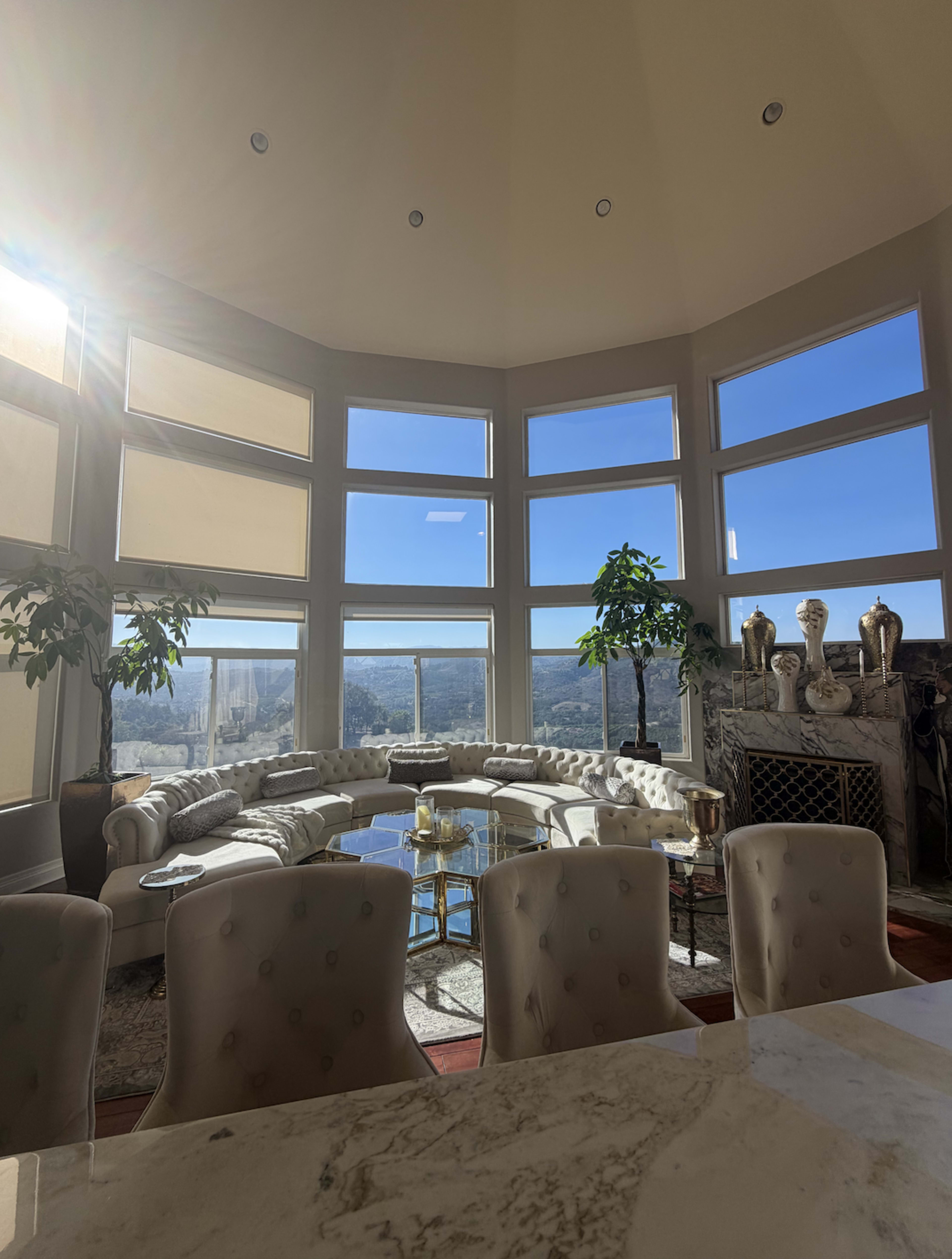 A spacious living room features large windows with a panoramic view, elegant furniture, and decorative plants.