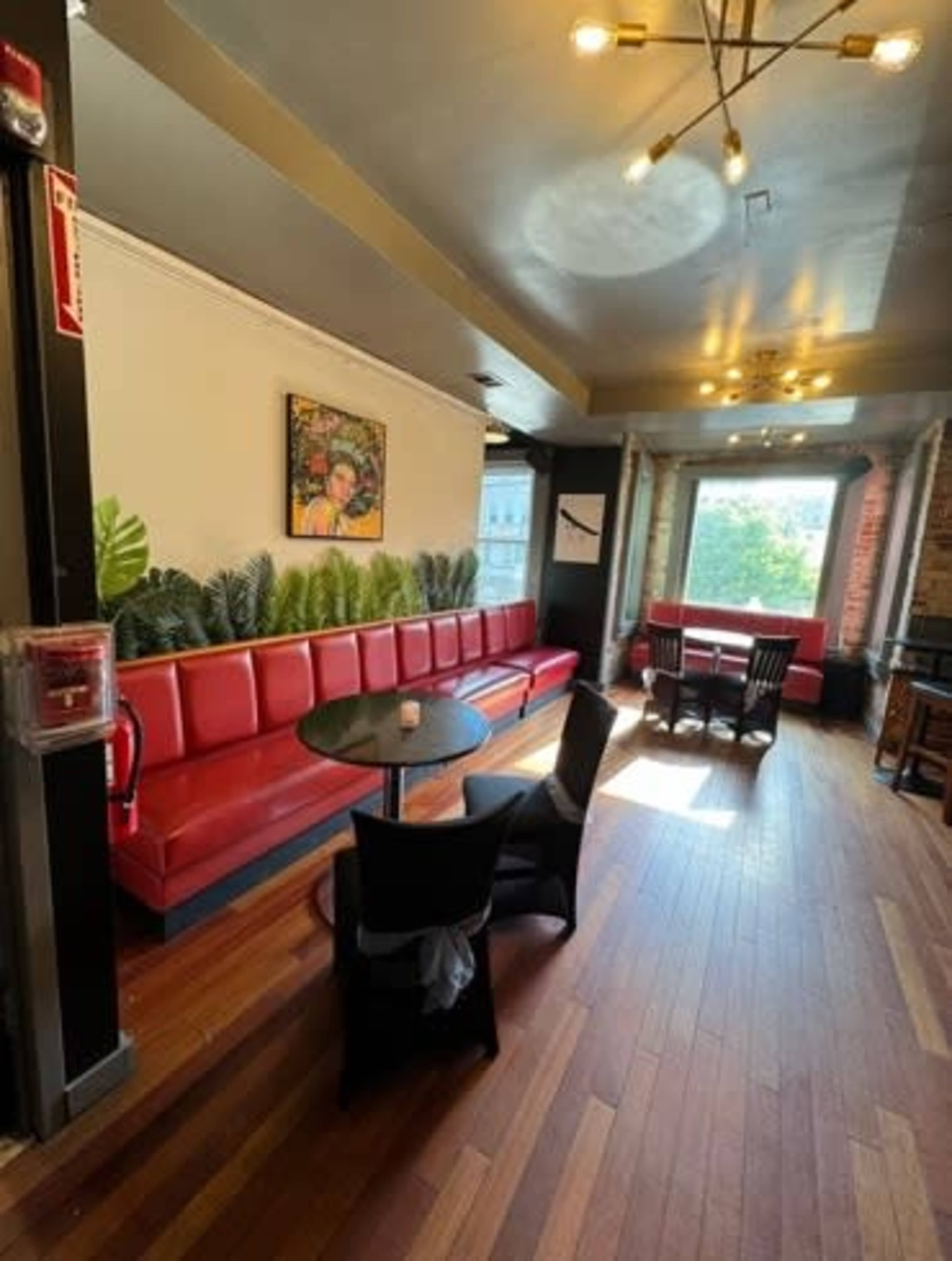 The image shows a cafe interior with red seating along the wall, a round table with black chairs, and large windows letting in natural light.