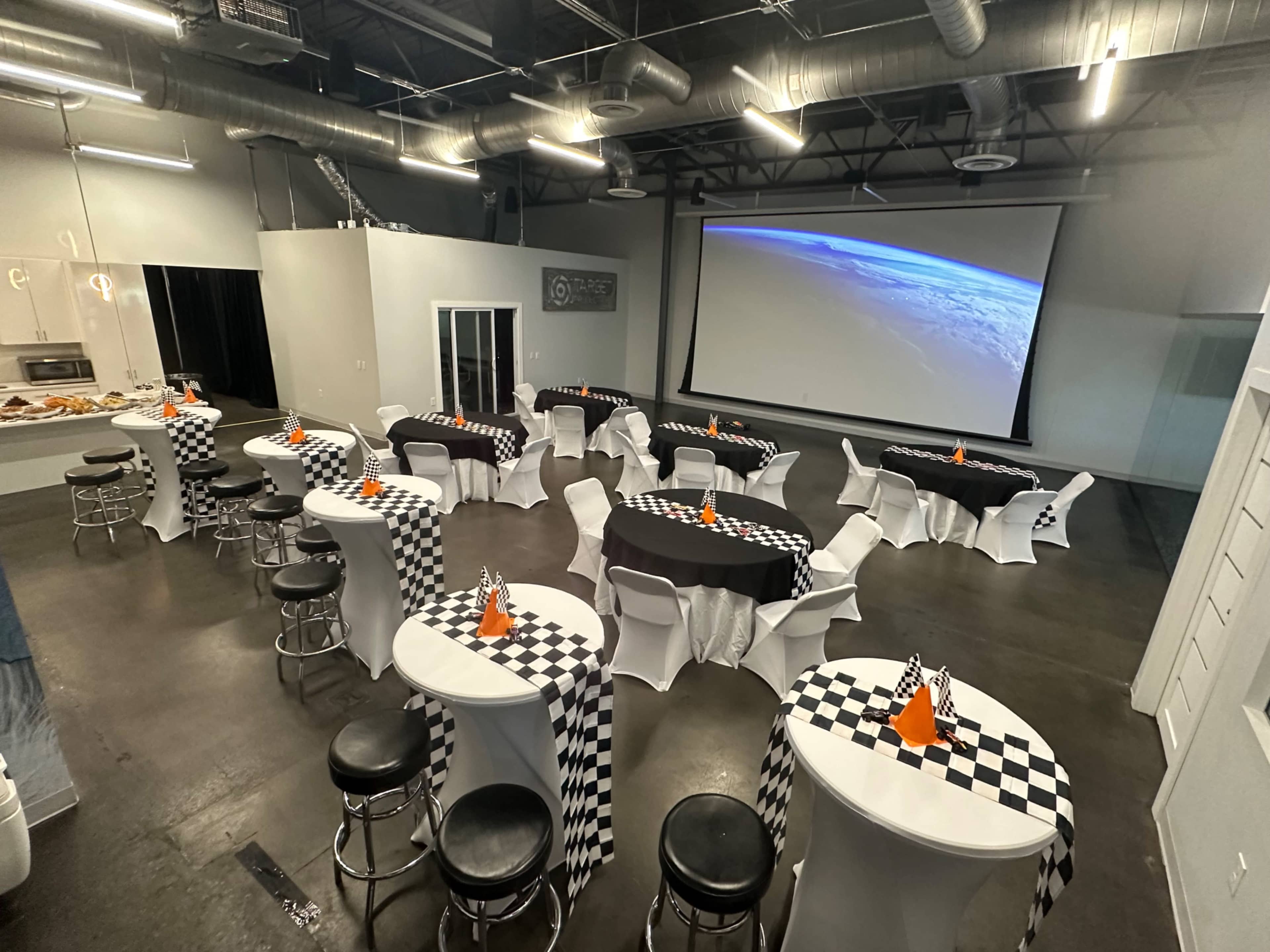 The image shows a spacious event room set up with checkered tablecloths and orange decorations on circular tables, with a large projection screen displaying an image of Earth.