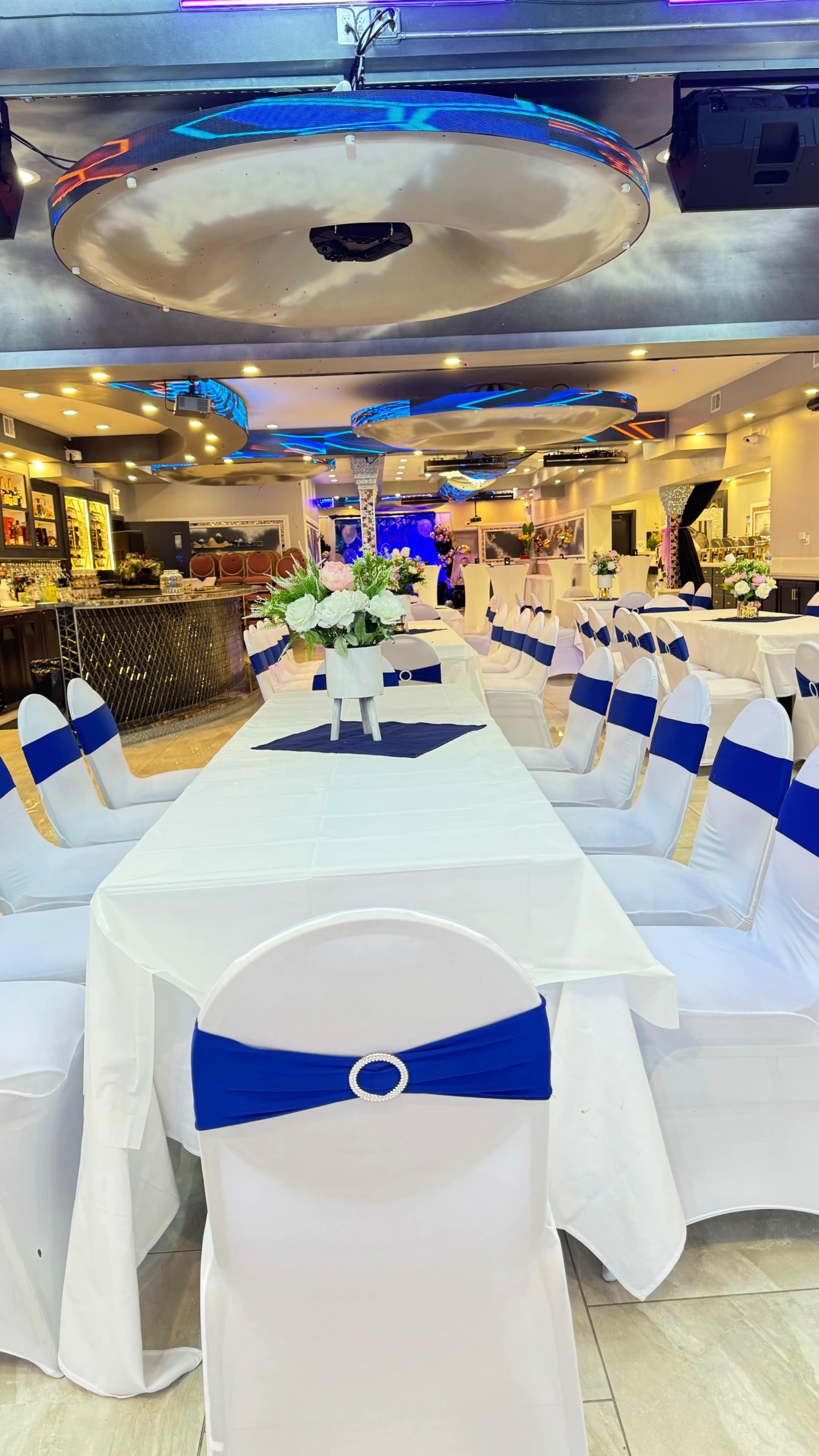 A spacious restaurant interior features a long table set with white tablecloths and blue-accented chair covers, surrounded by decorative flowers and ambient lighting.