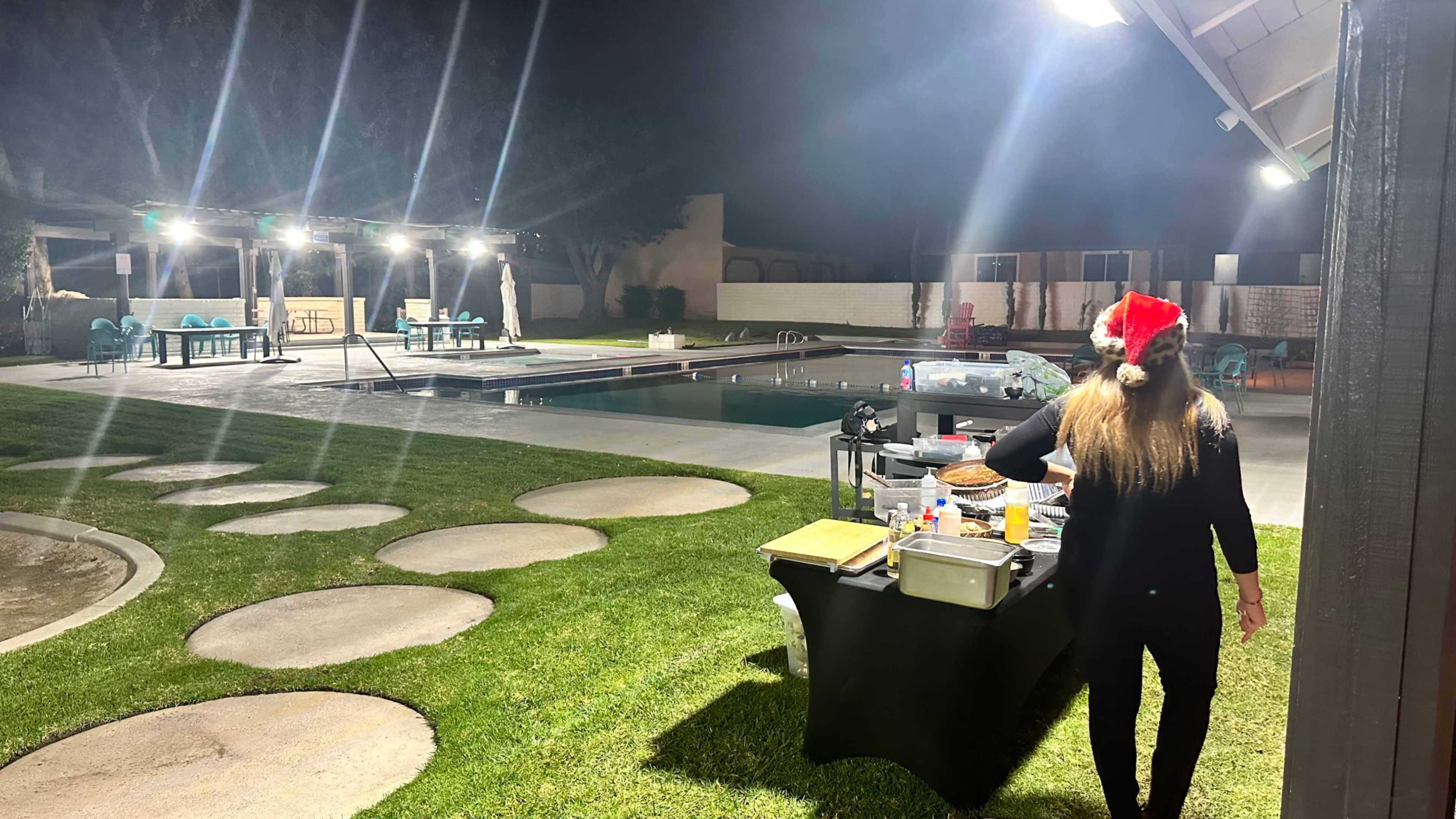 A person wearing a Christmas hat stands by a table with food, overlooking a pool area illuminated by string lights at night.