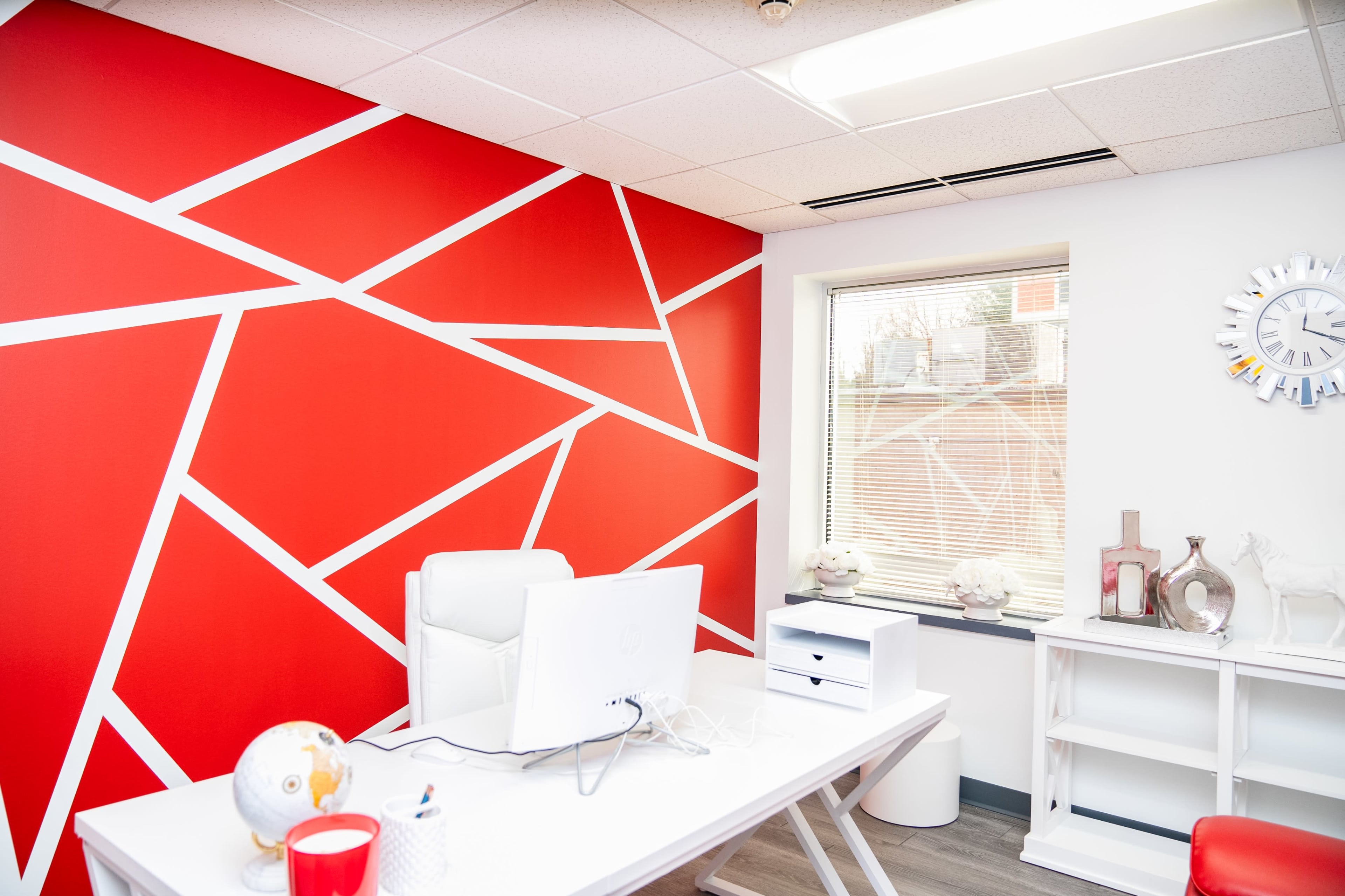 A modern office features a red accent wall with geometric white lines, a white desk with a computer, and minimalist decor including a globe and a clock.