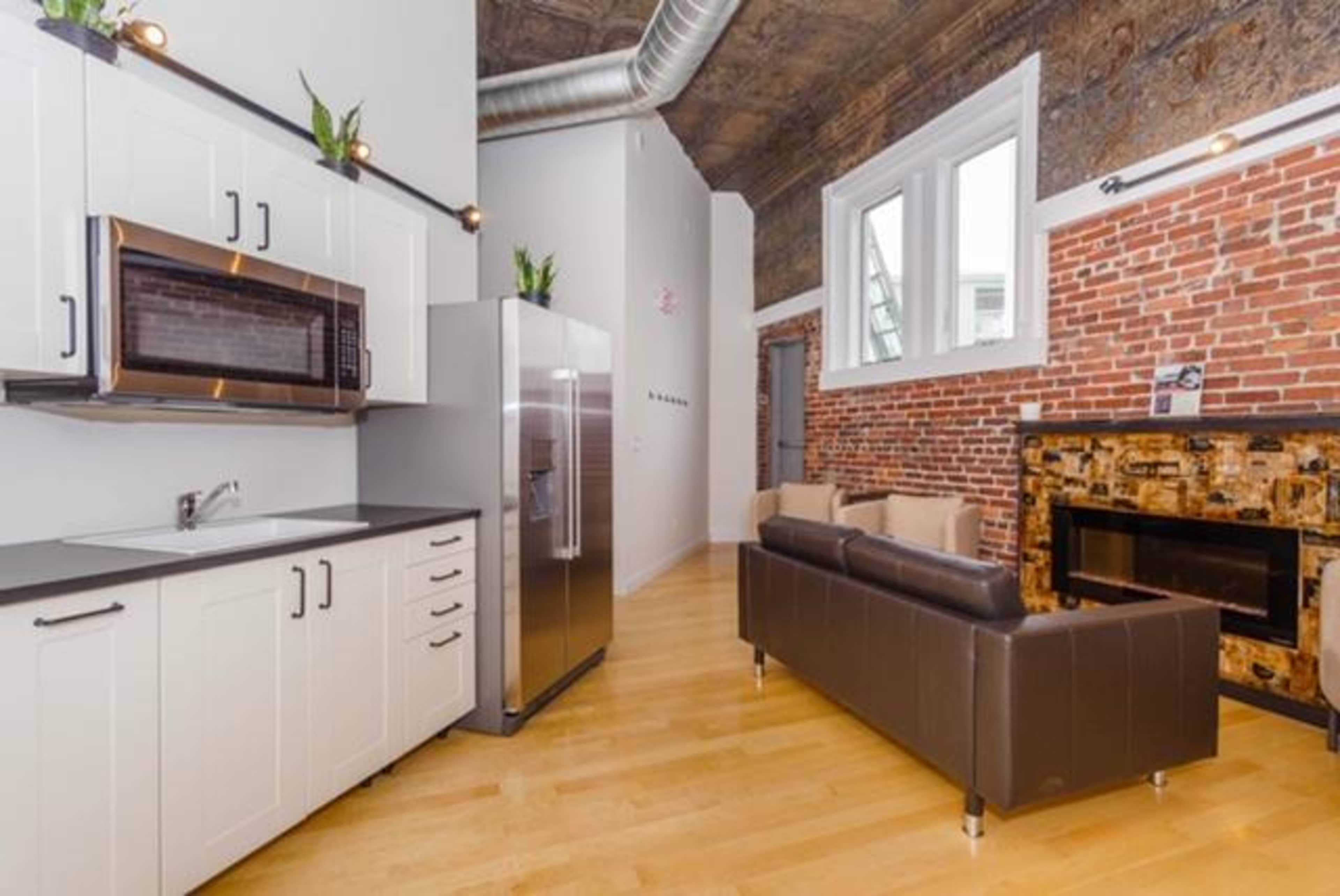 A modern kitchen with white cabinetry and stainless steel appliances, adjoining a living area with a couch and a decorative fireplace, set against exposed brick walls.