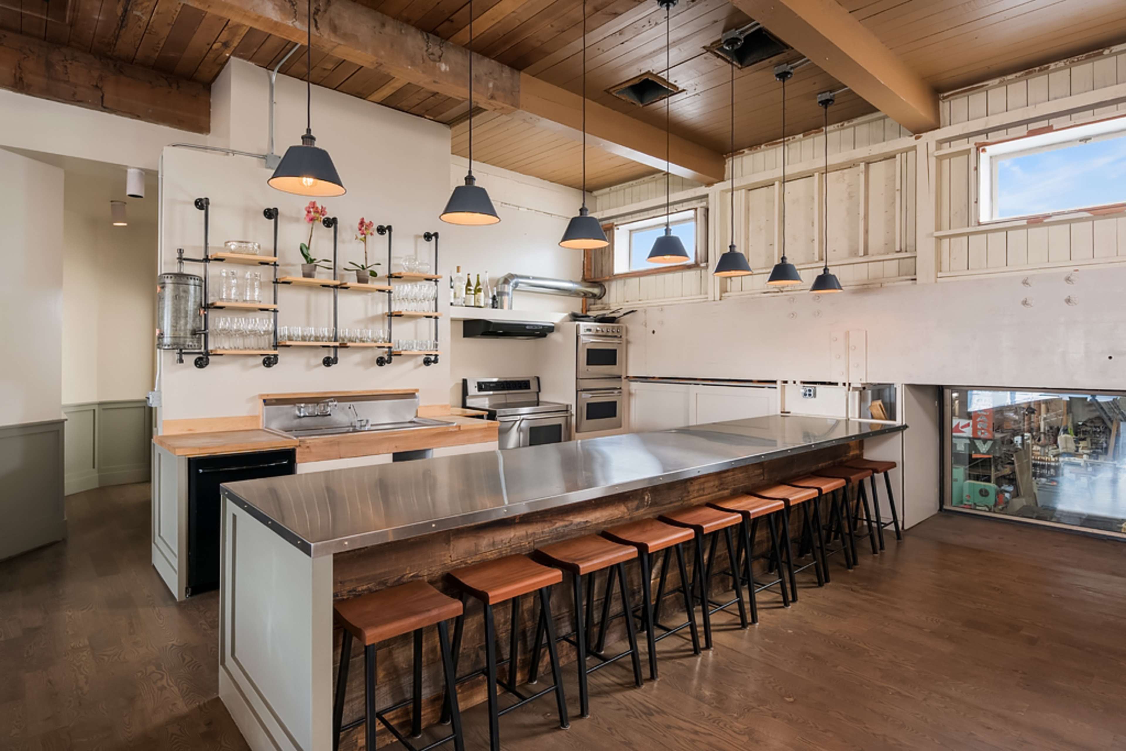 A modern kitchen with a stainless steel countertop, several hanging pendant lights, and a row of wooden bar stools.